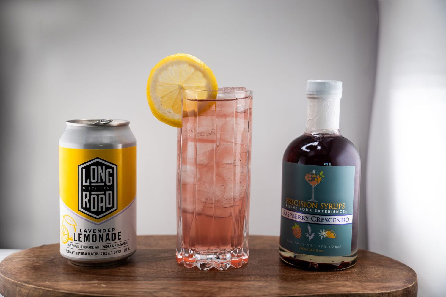 Refreshing pink drink with lemon garnish displayed between canned lemonade and artisanal syrup bottle.