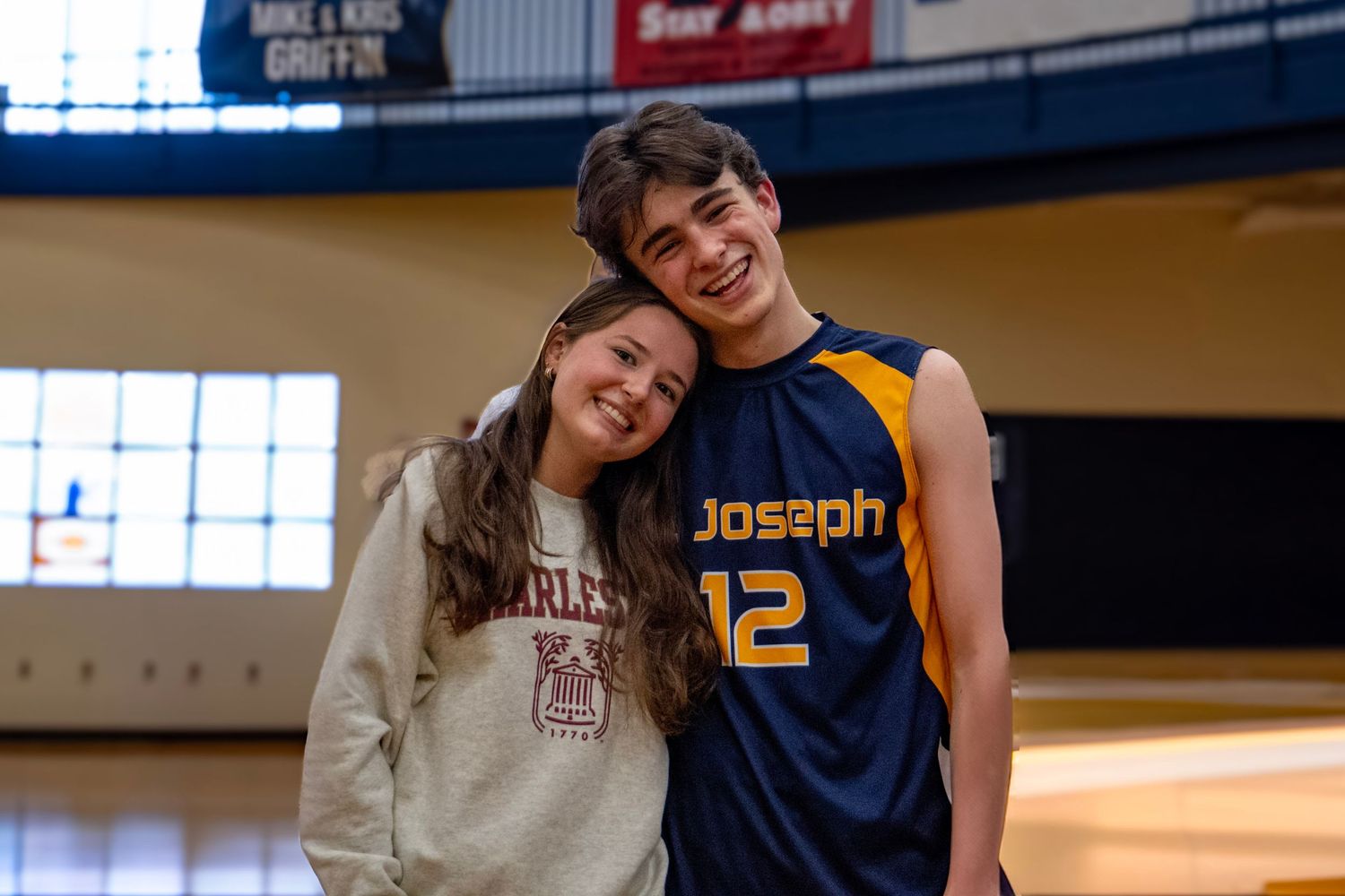 Two students posing together in a school gym with athletic banners visible.