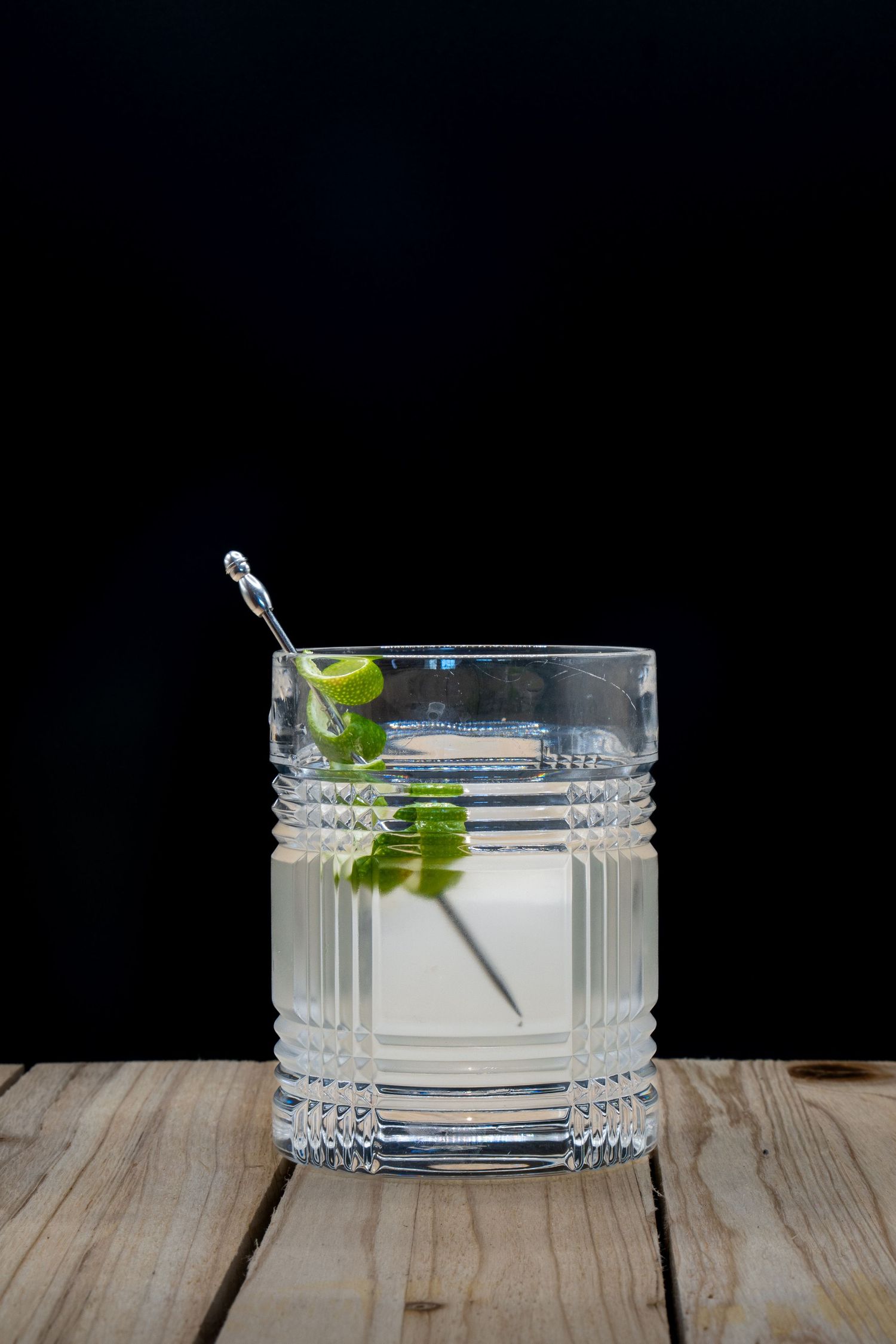 Clear cocktail in textured crystal glass with lime and metal stirrer on wooden surface.