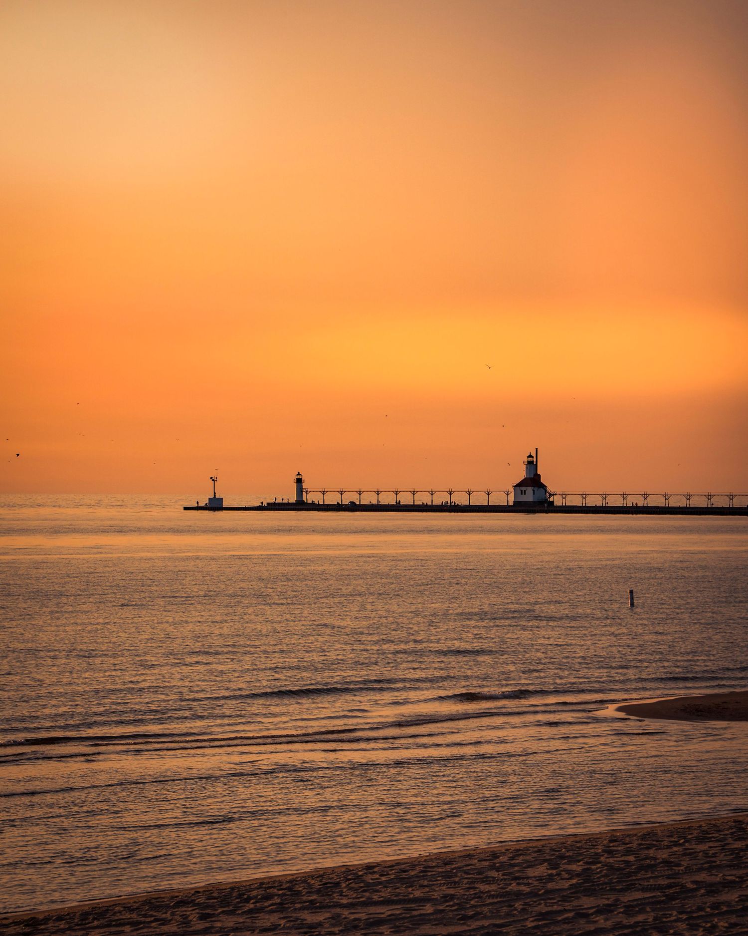 Lighthouse and pier silhouetted against a dramatic orange sunset sky over calm waters.