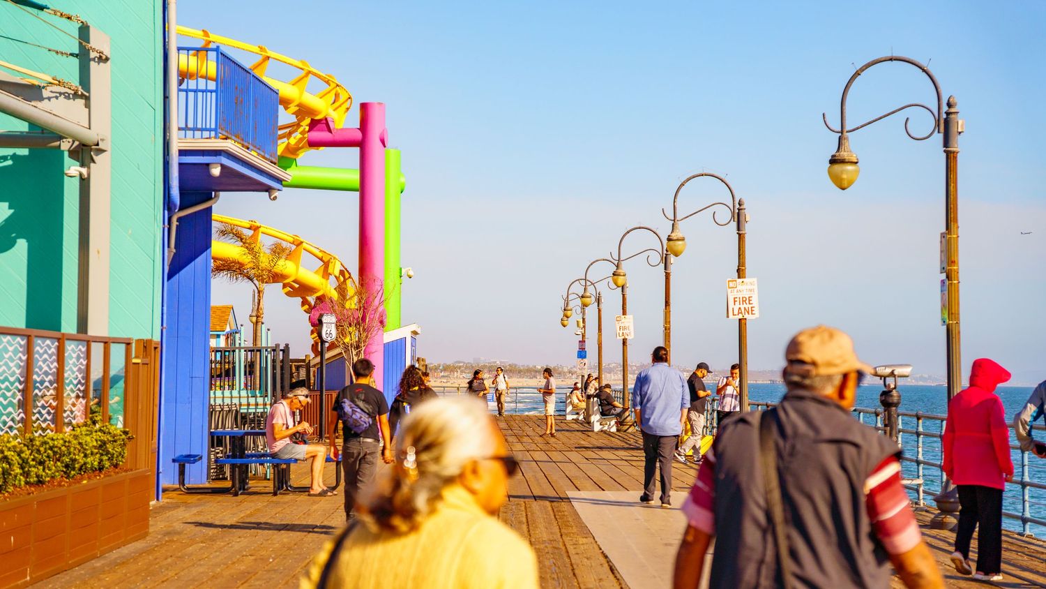 Colorful boardwalk entrance with vintage lampposts and people walking at Pacific Park during golden hour.