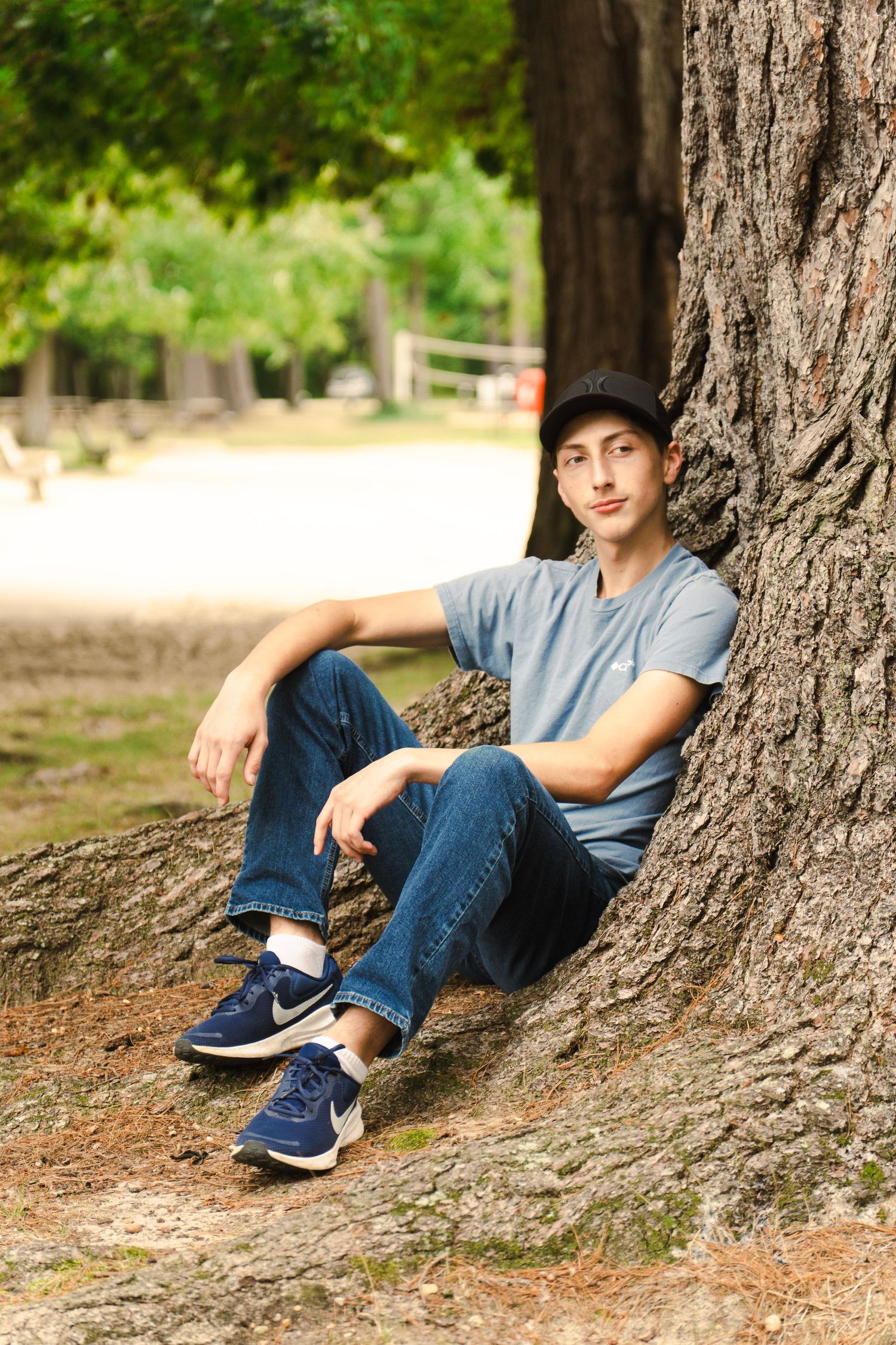 Young man in a gray t-shirt, jeans and blue sneakers sits relaxed against a large tree trunk in a sunny park.