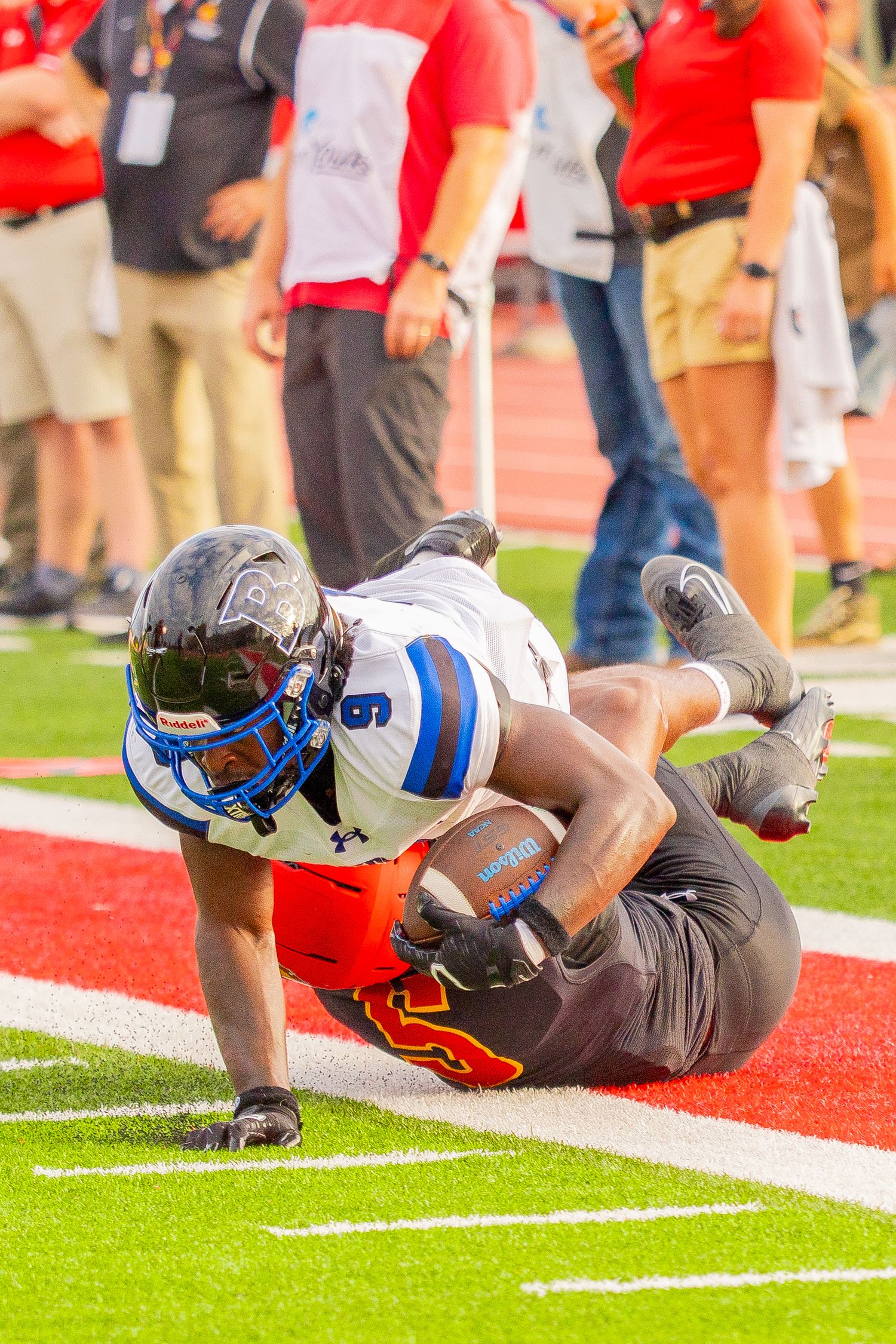 Football player in a white and blue number 9 jersey dives with the ball across the sideline as sideline staff watch.