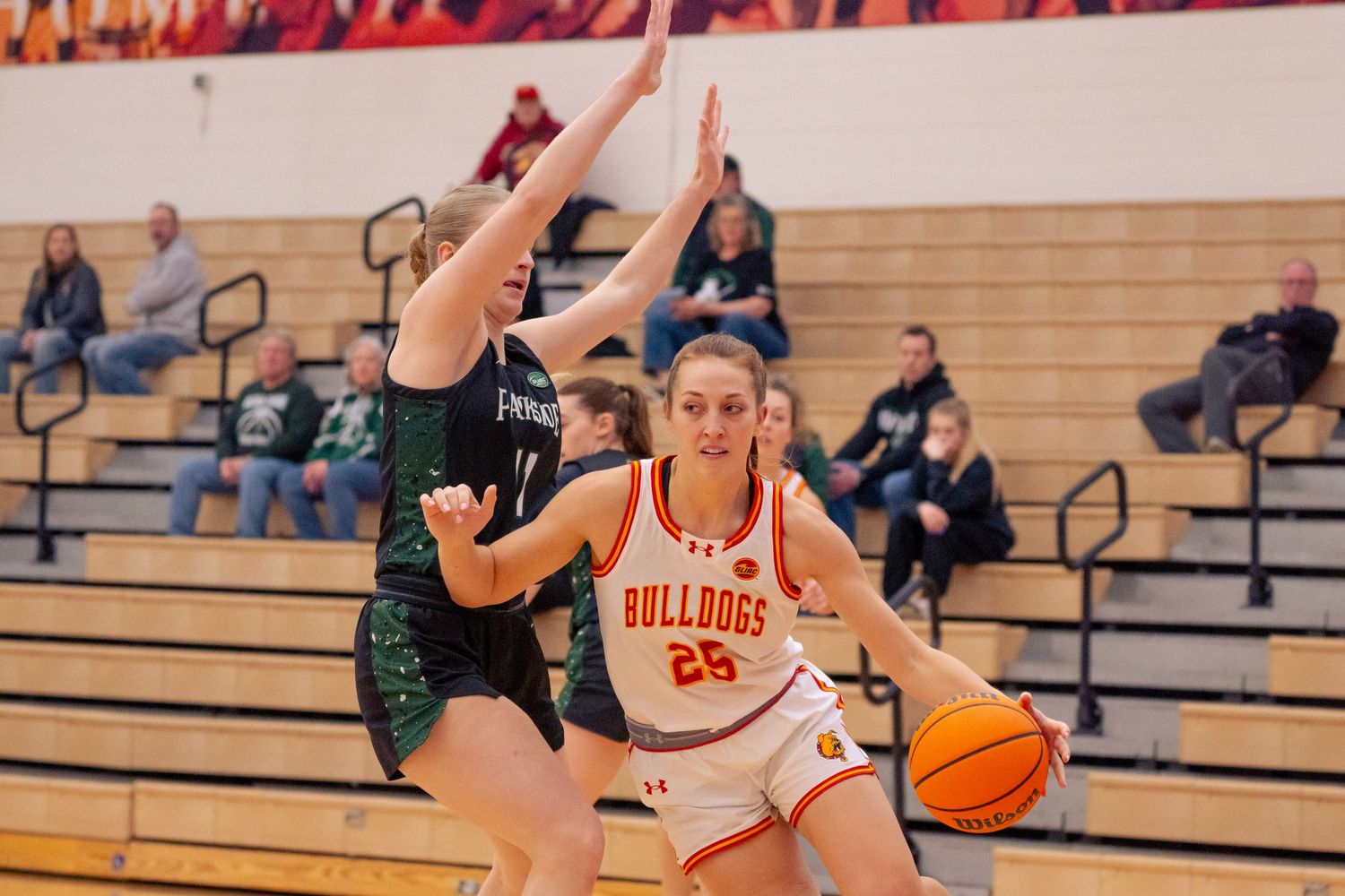 Bulldogs number 25 basketball player in white and orange dribbles past a defender in green as spectators watch from the bleachers.
