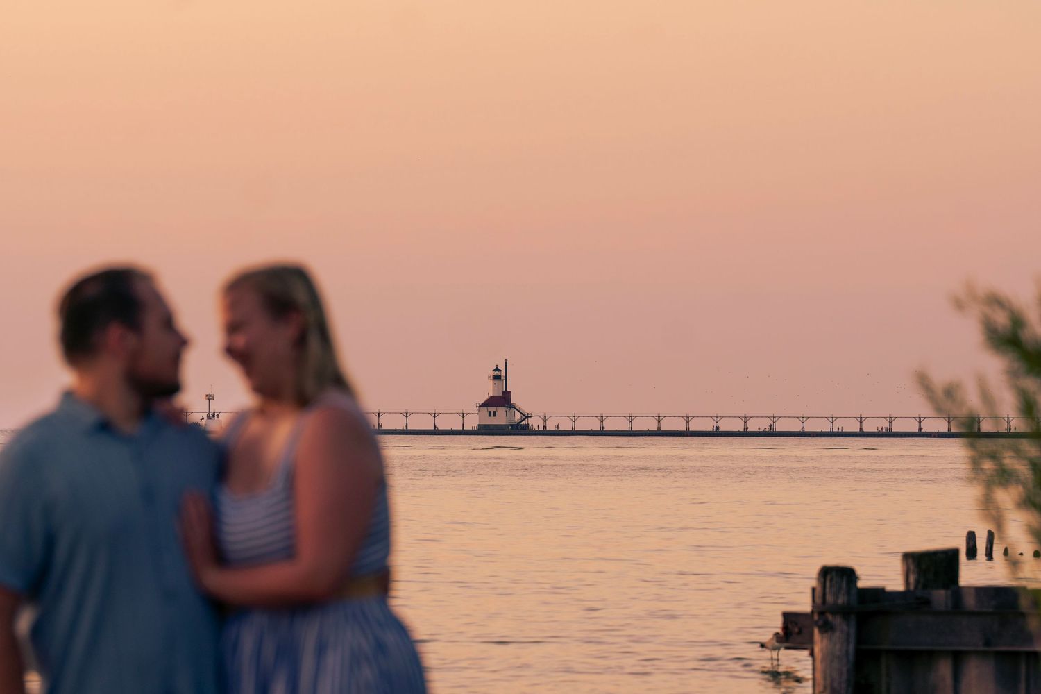 Silhouetted couple embraces on beach at sunset with pier in background as orange sky glows over ocean.