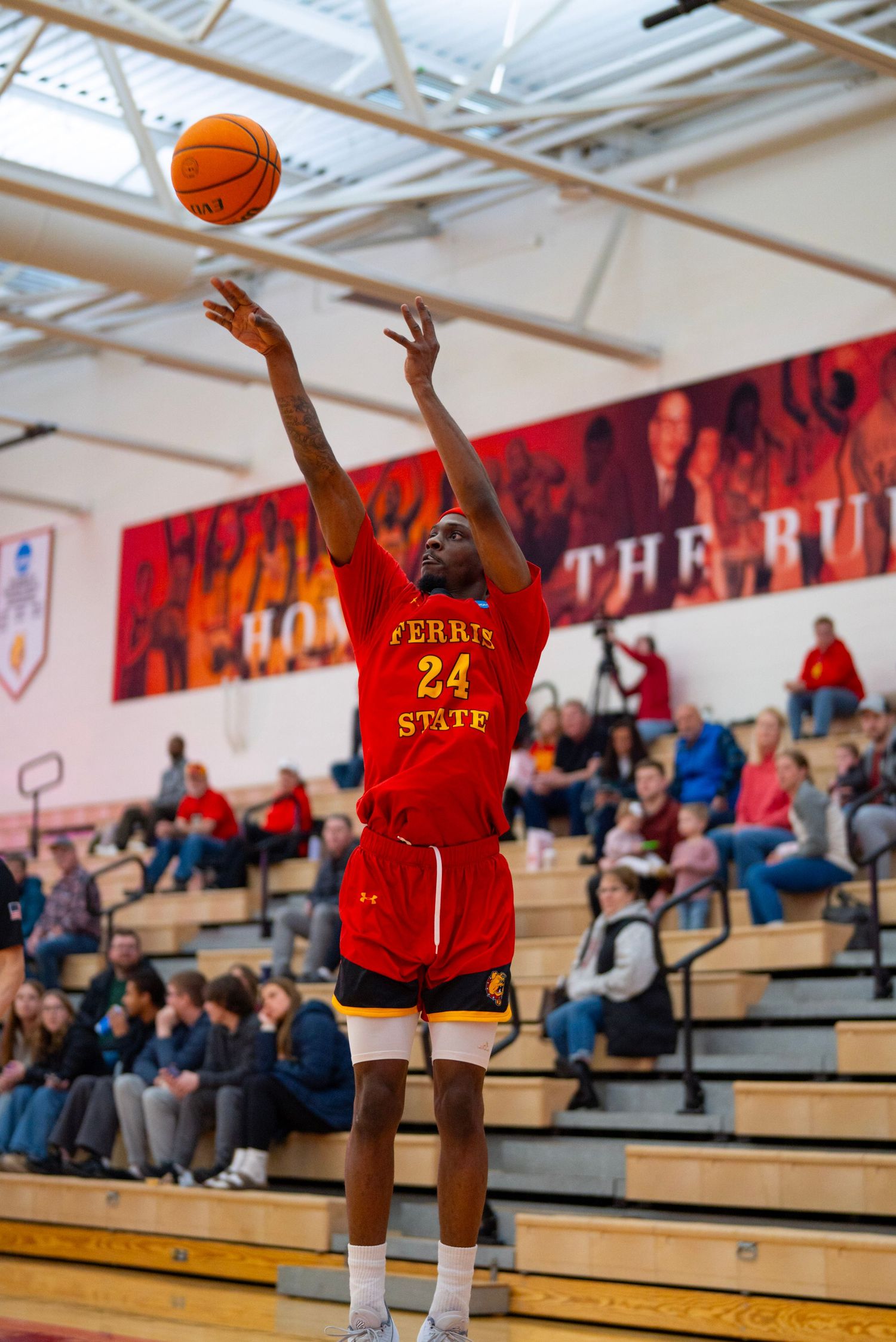 Basketball player in red jersey jumps to shoot the ball during indoor game while spectators watch from bleachers.