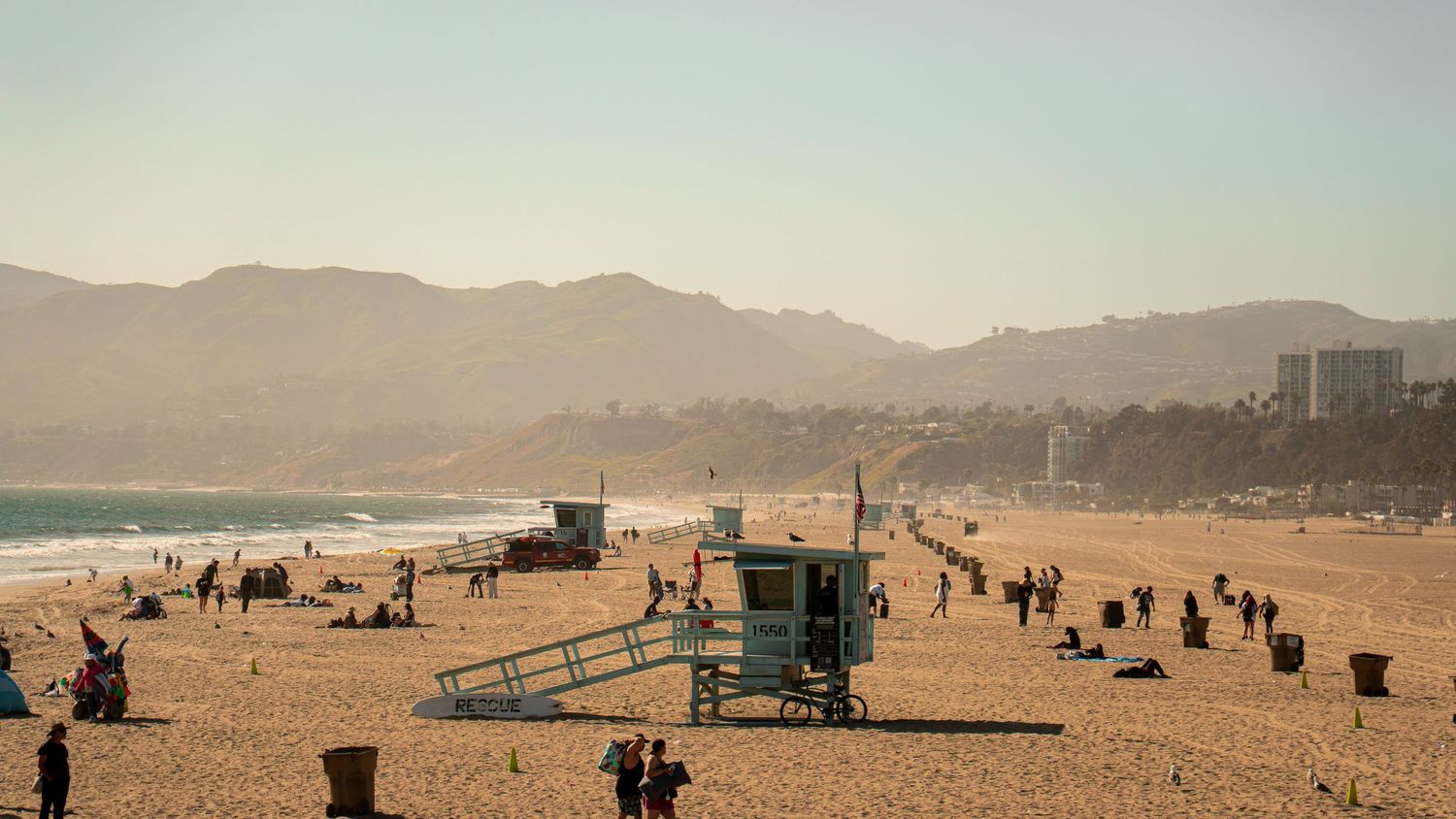 Wide angle view of a busy beach with lifeguard stations and mountains in the background.