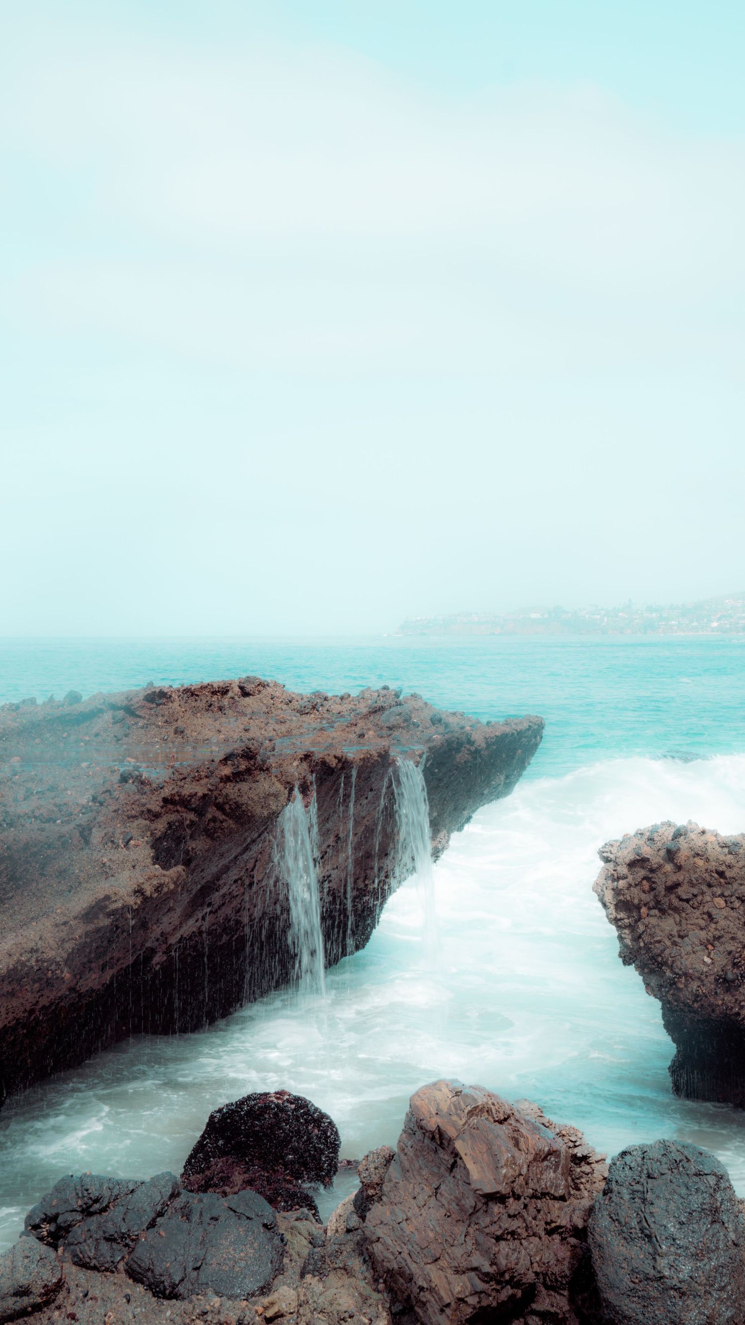 Small waterfall flows between rocky coastal formations into turquoise waters below.