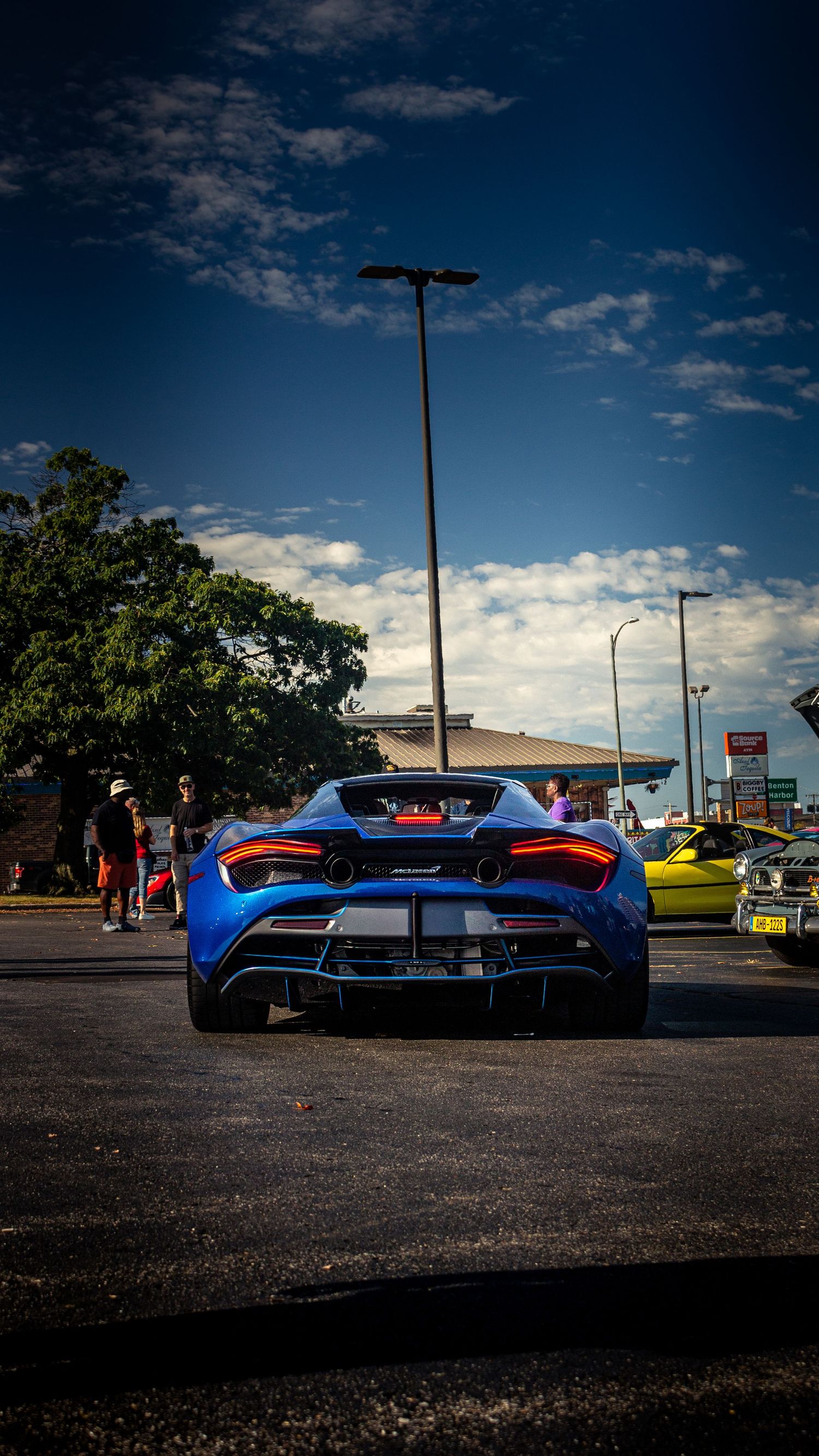 Rear view of blue McLaren 720S in parking lot under dramatic sky.