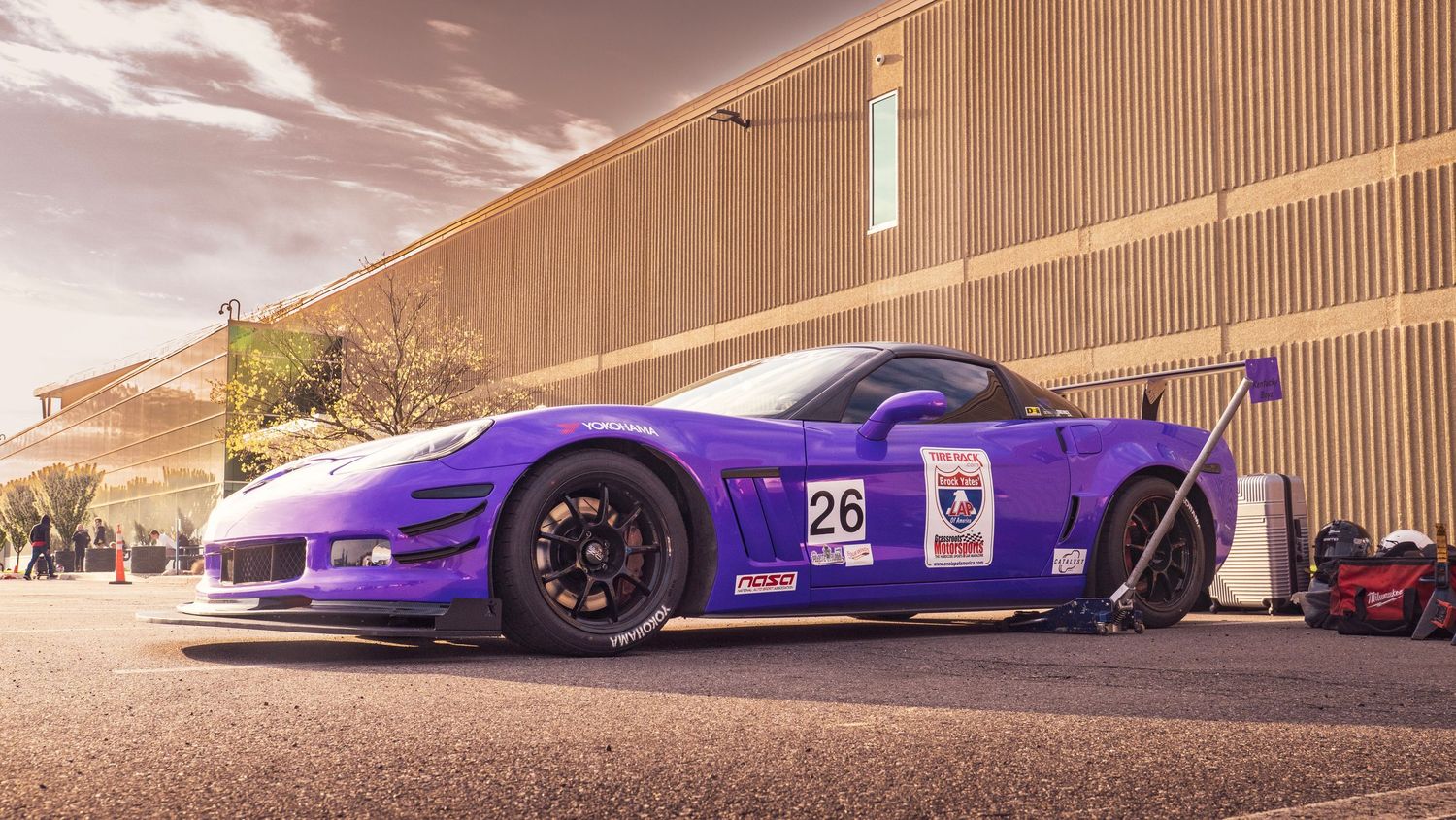 Purple sports car silhouette against sunset with wooden building facade in background.