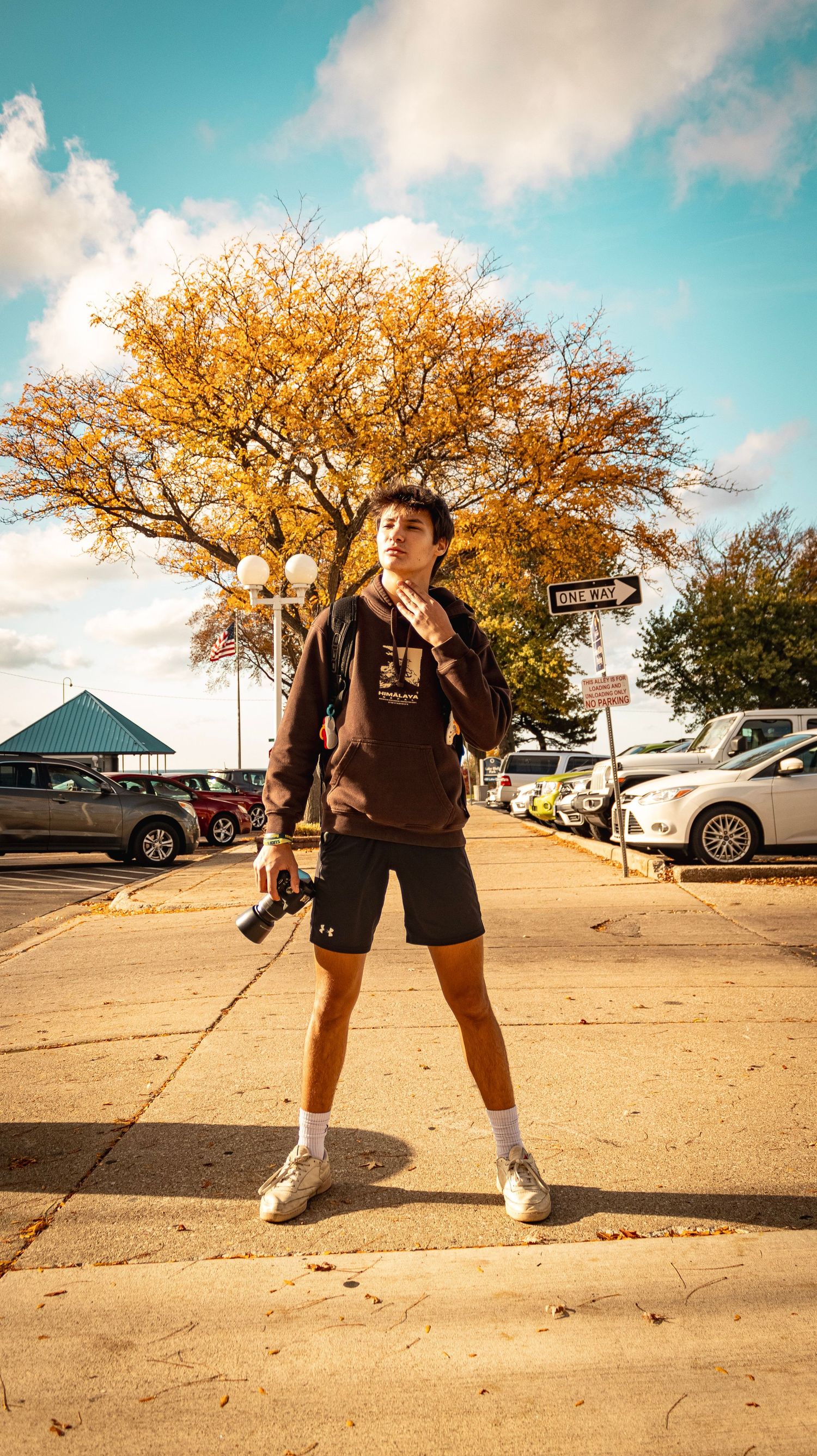 Person holding camera poses under golden autumn tree in parking lot.