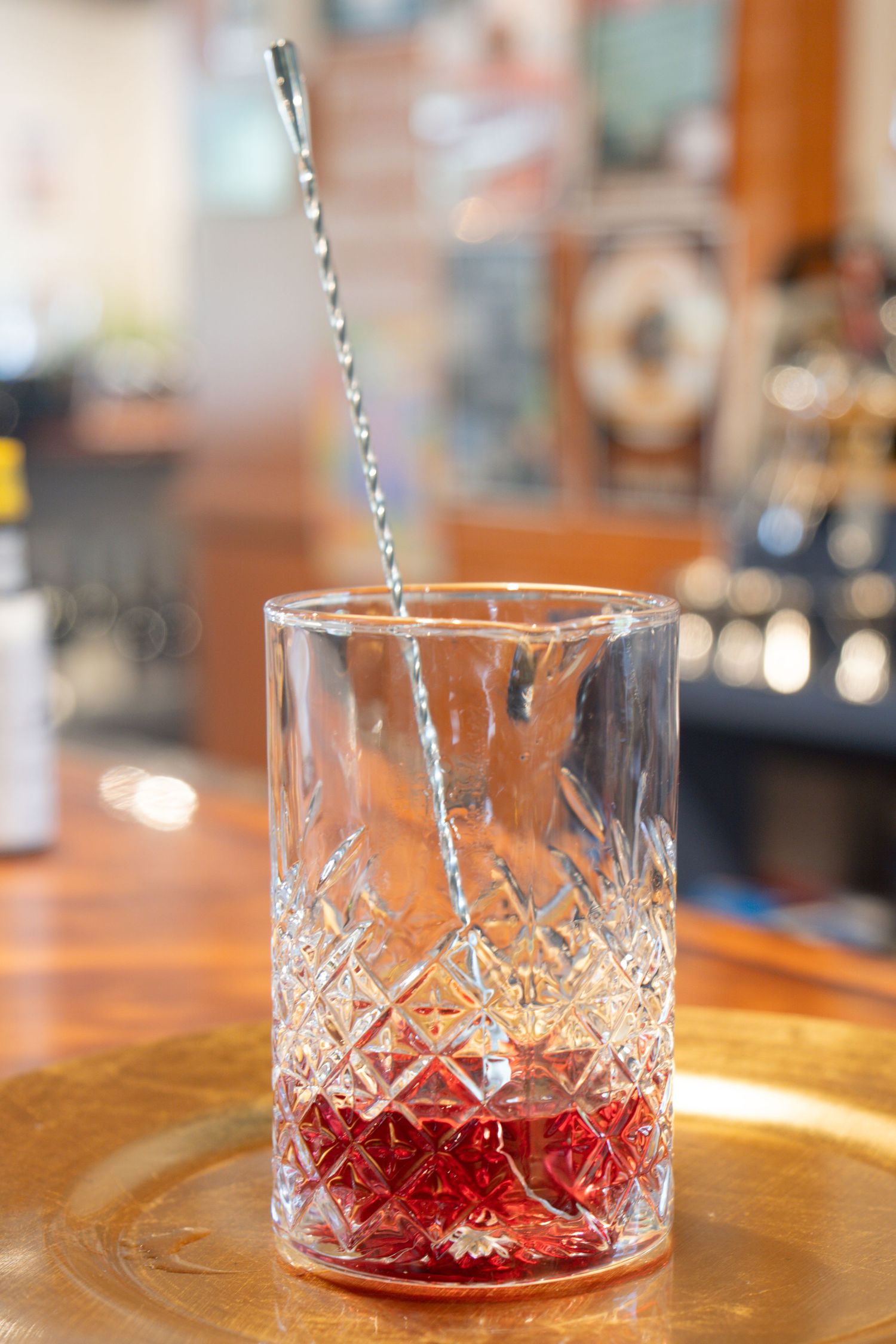 Crystal mixing glass with red cocktail syrup and striped straw in bar setting.