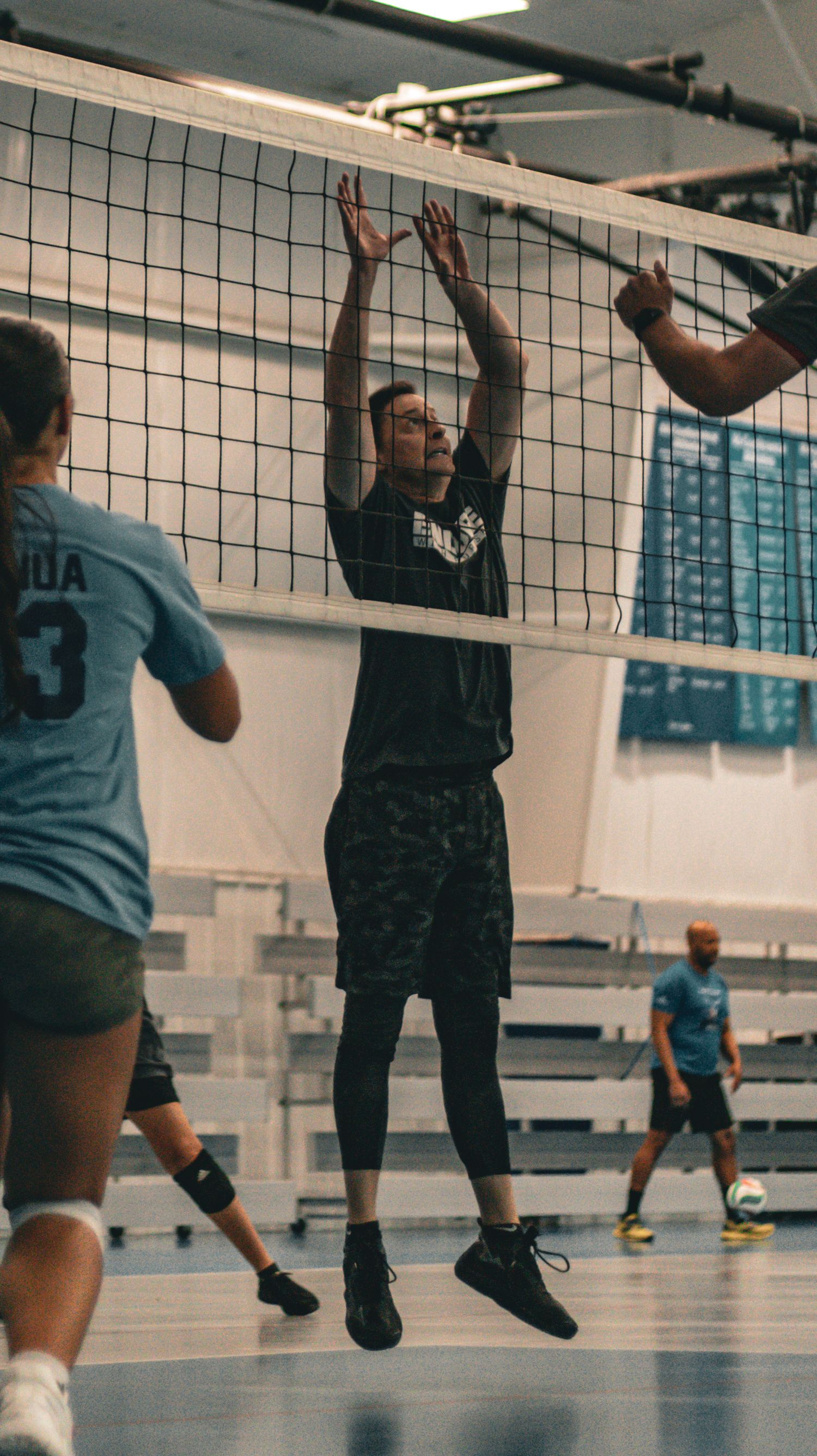 Volleyball player jumps to block at net during indoor game with opponents preparing to spike.