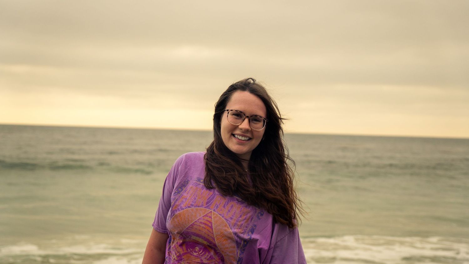 Person wearing purple graphic tee stands on beach during sunset with calm ocean behind.