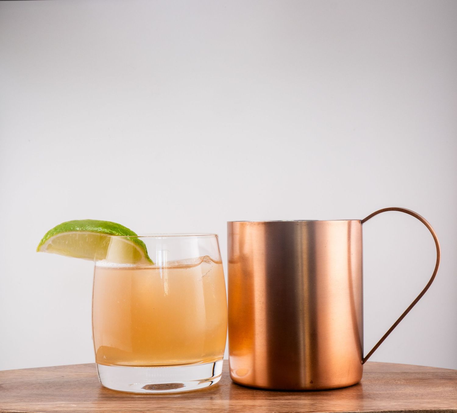 Refreshing cocktail with lime wedge next to copper mug on wooden surface.