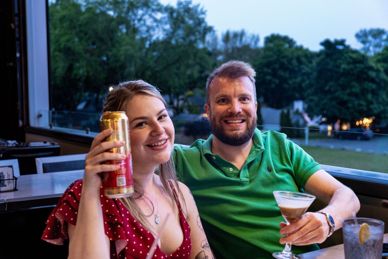 Two people enjoying drinks together outdoors at dusk with trees visible in the background.