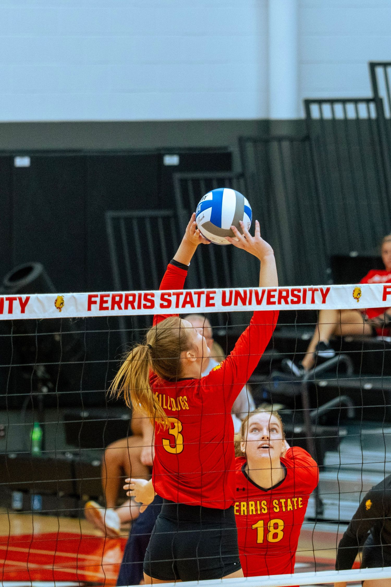 Volleyball players in red uniforms leap to block a shot at the net during a Ferris State University match.