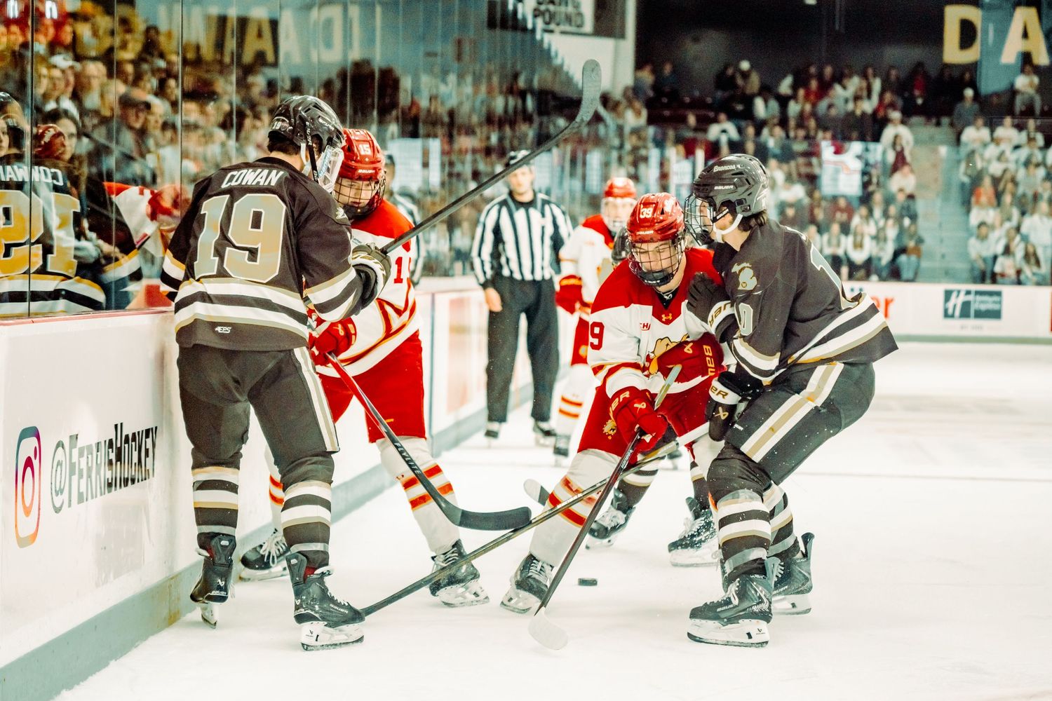 Hockey players in dark and red-and-white jerseys battle for the puck along the boards during an ice hockey game as a referee looks on.