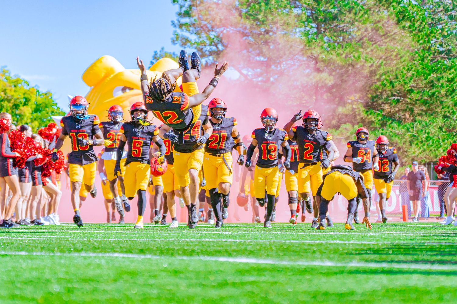 Football players in black and gold uniforms with red helmets run onto a grass field through orange smoke as a teammate is lifted overhead.