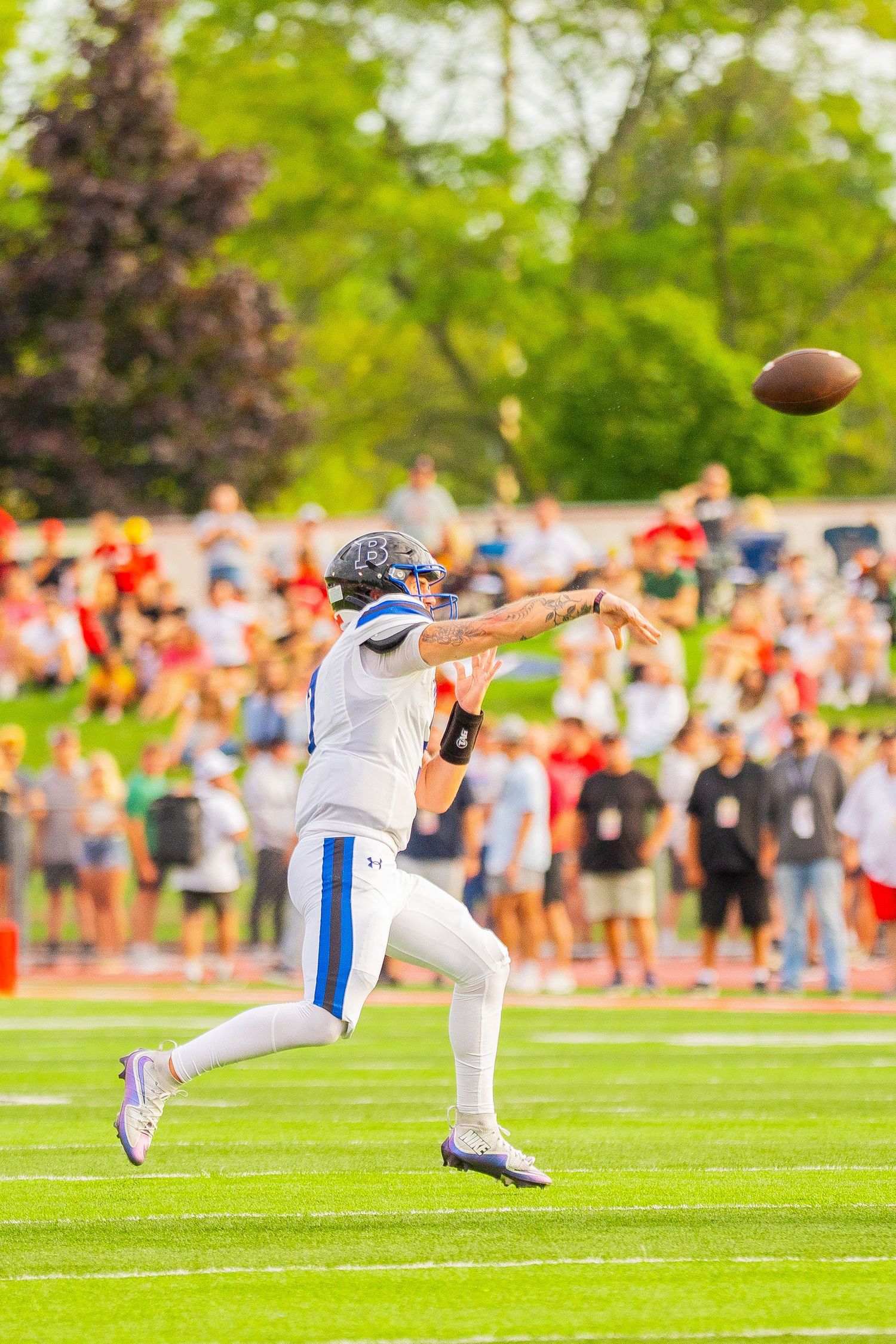 Football quarterback in a white and blue uniform and helmet throws a pass during a game while spectators watch from the sidelines.