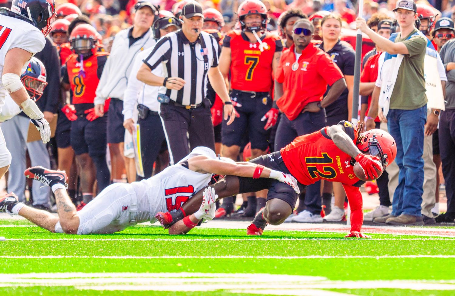 Football player in red jersey number 12 tackles a ball carrier in white along the sideline as a referee and crowd look on.