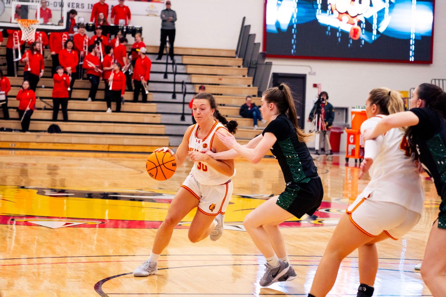 Women's basketball player in a white uniform dribbles past a defender in green as red-clad teammates watch from the bleachers.