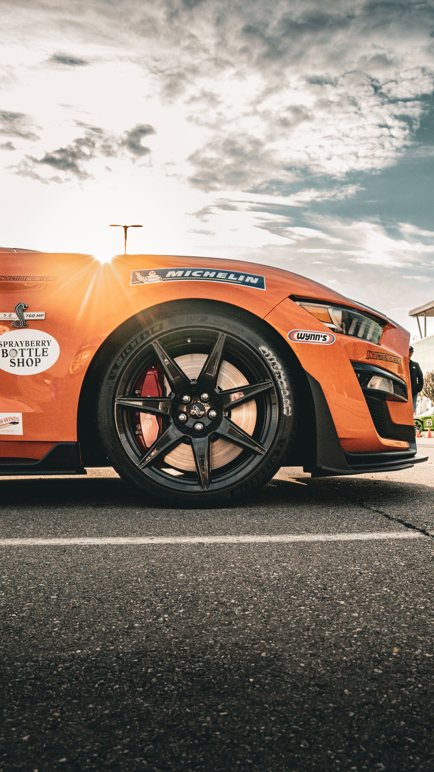 Low angle view of an orange Mustang Shelby GT500 with Michelin tires and black wheels against cloudy sky.