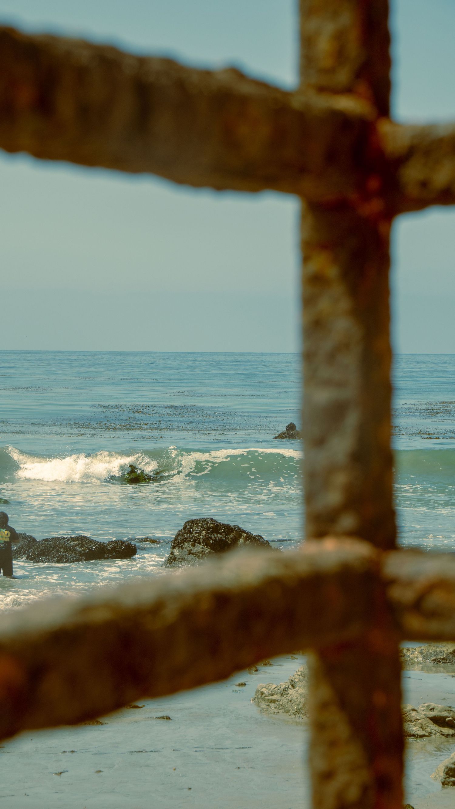 Ocean waves viewed through rustic metal fence bars along rocky coastline.