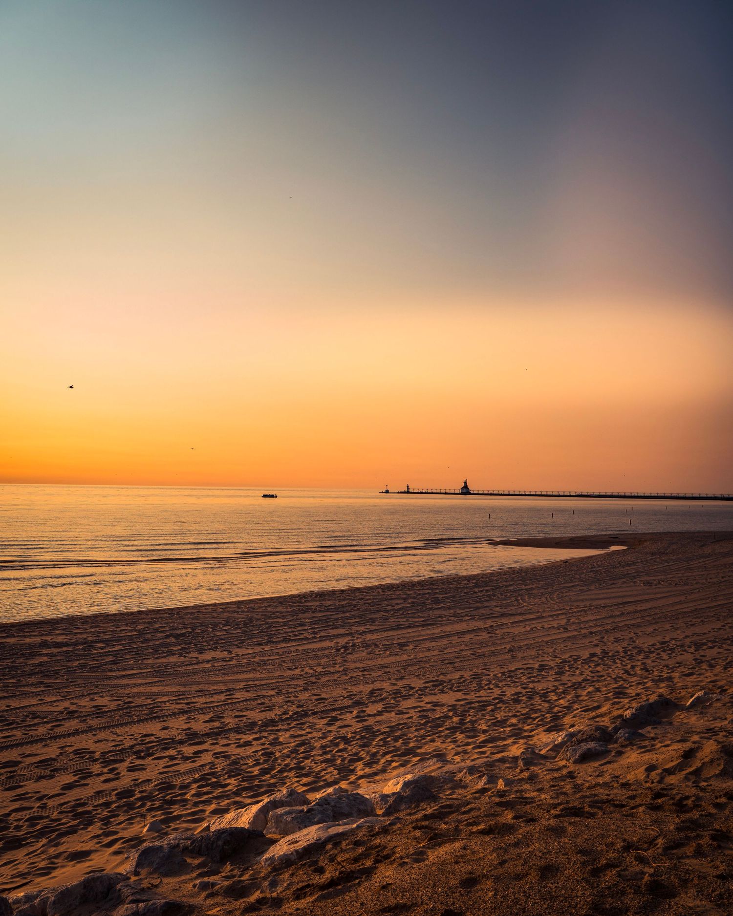 Tranquil beach sunset with distant lighthouse silhouette and golden reflections on water.