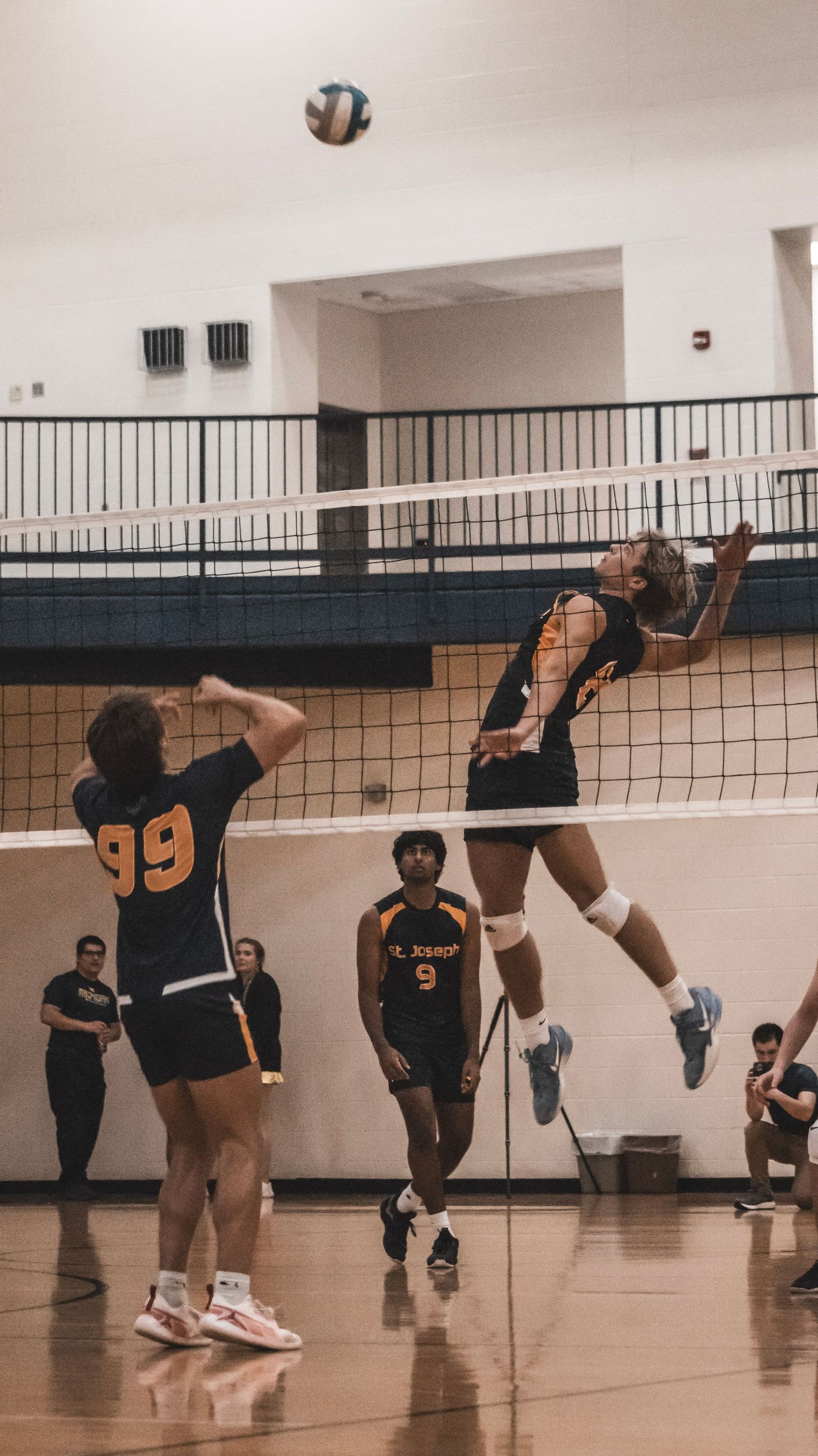 Player leaps high to spike volleyball while teammate watches during indoor match.