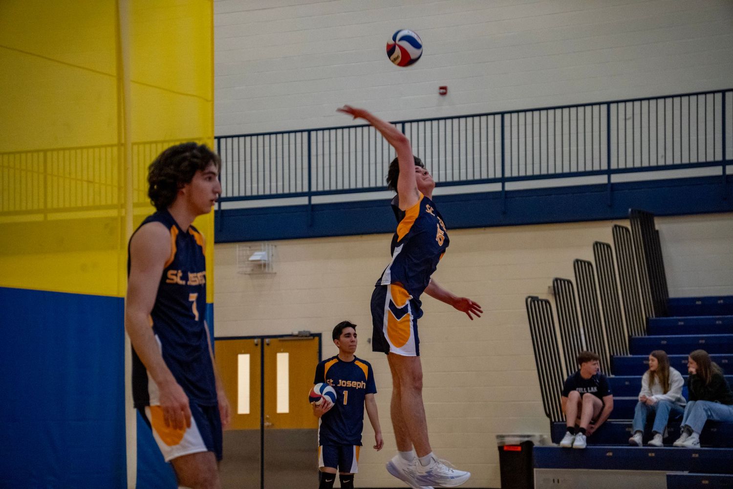 Players in navy and yellow uniforms compete for a volleyball in a gymnasium setting.