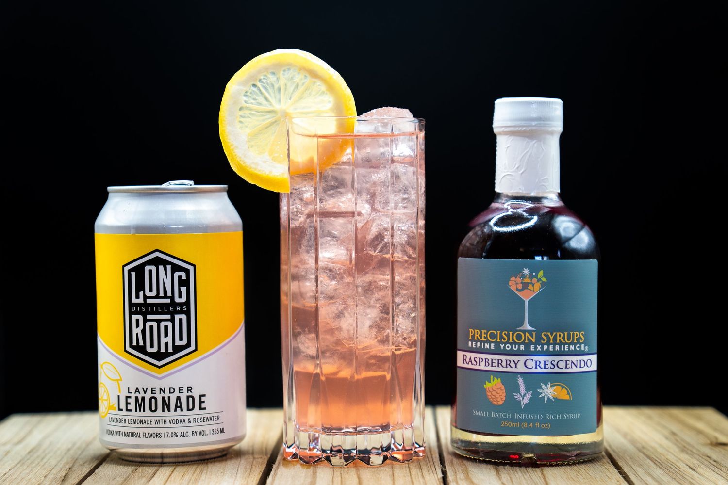 Craft beverage display featuring lemonade can, pink cocktail, and premium spirits bottle on wood surface.