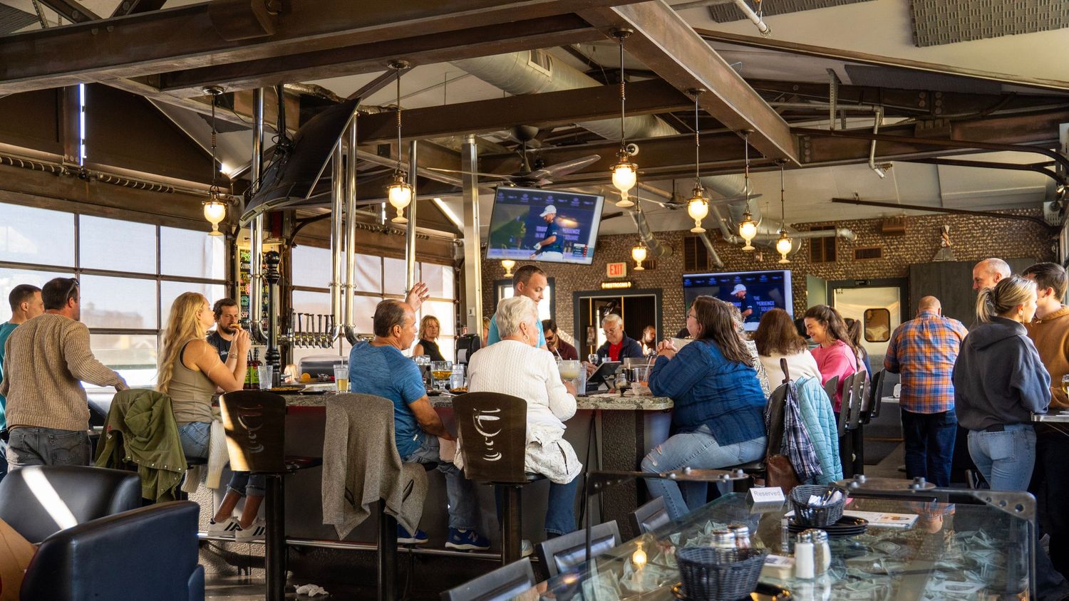Customers enjoy drinks at a busy industrial-style bar with exposed ceiling beams and large windows during peak hours.