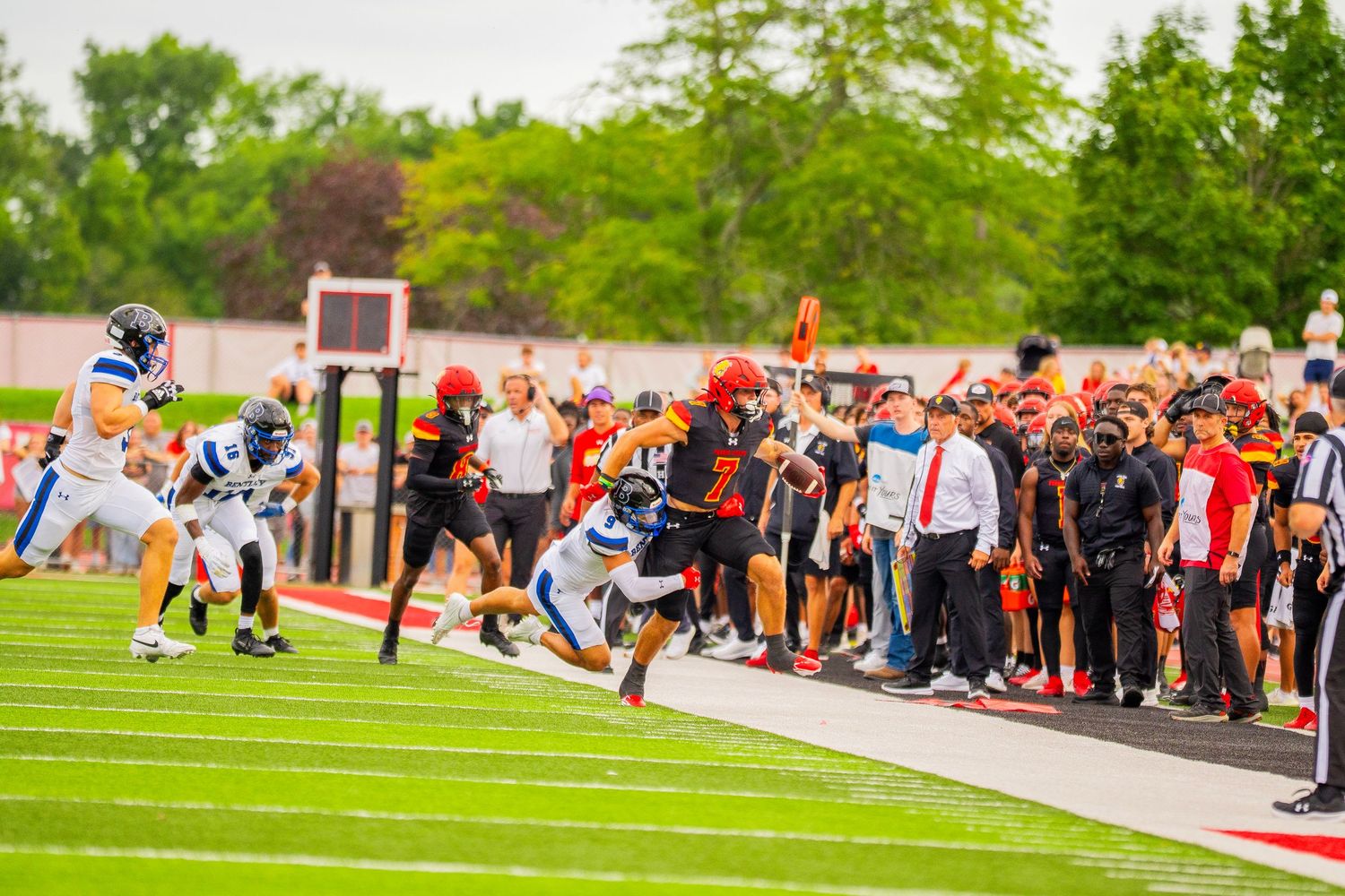 A football player in a white uniform races down the sideline past defenders as coaches and teammates in red and black watch the game.