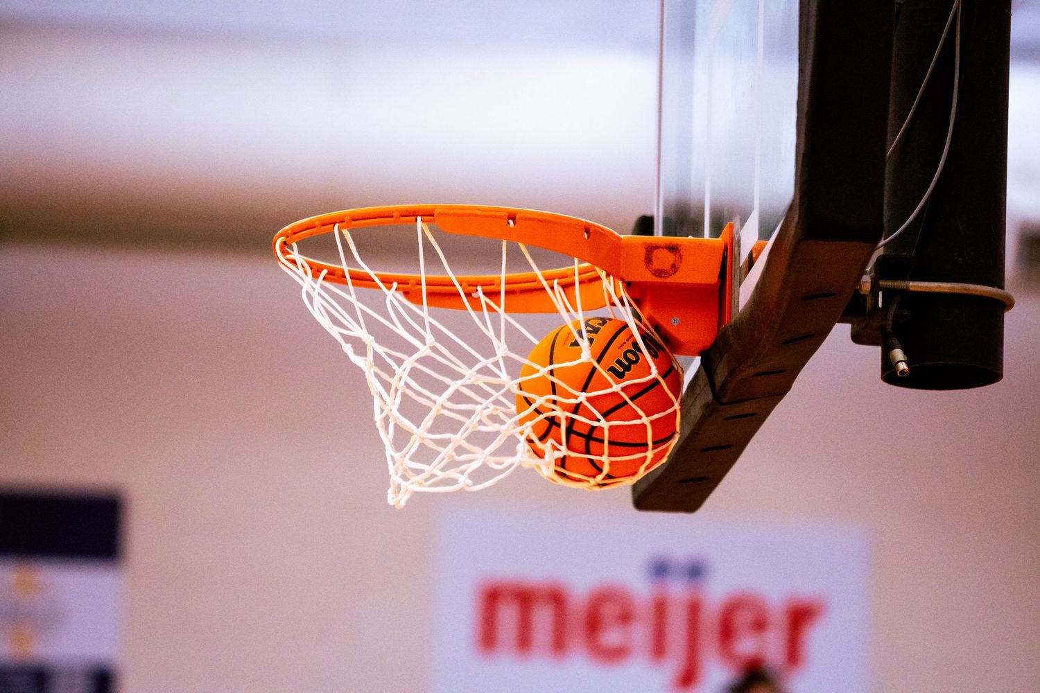 Basketball dropping through an orange hoop and white net at an indoor gym, with a Meijer banner on the wall in the blurred background.