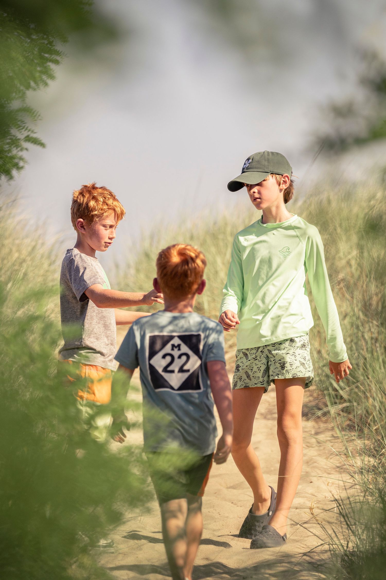 Three children walk together on a dusty trail during summer, framed through green foliage.
