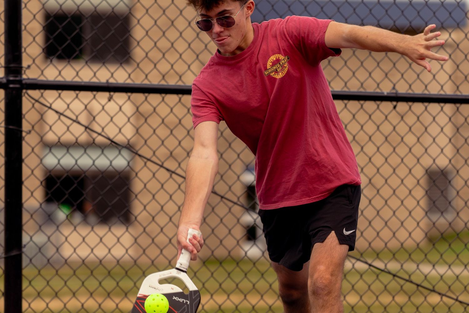 Player dressed in red returns a tennis ball during a match on a sunny day near a chain-link fence.