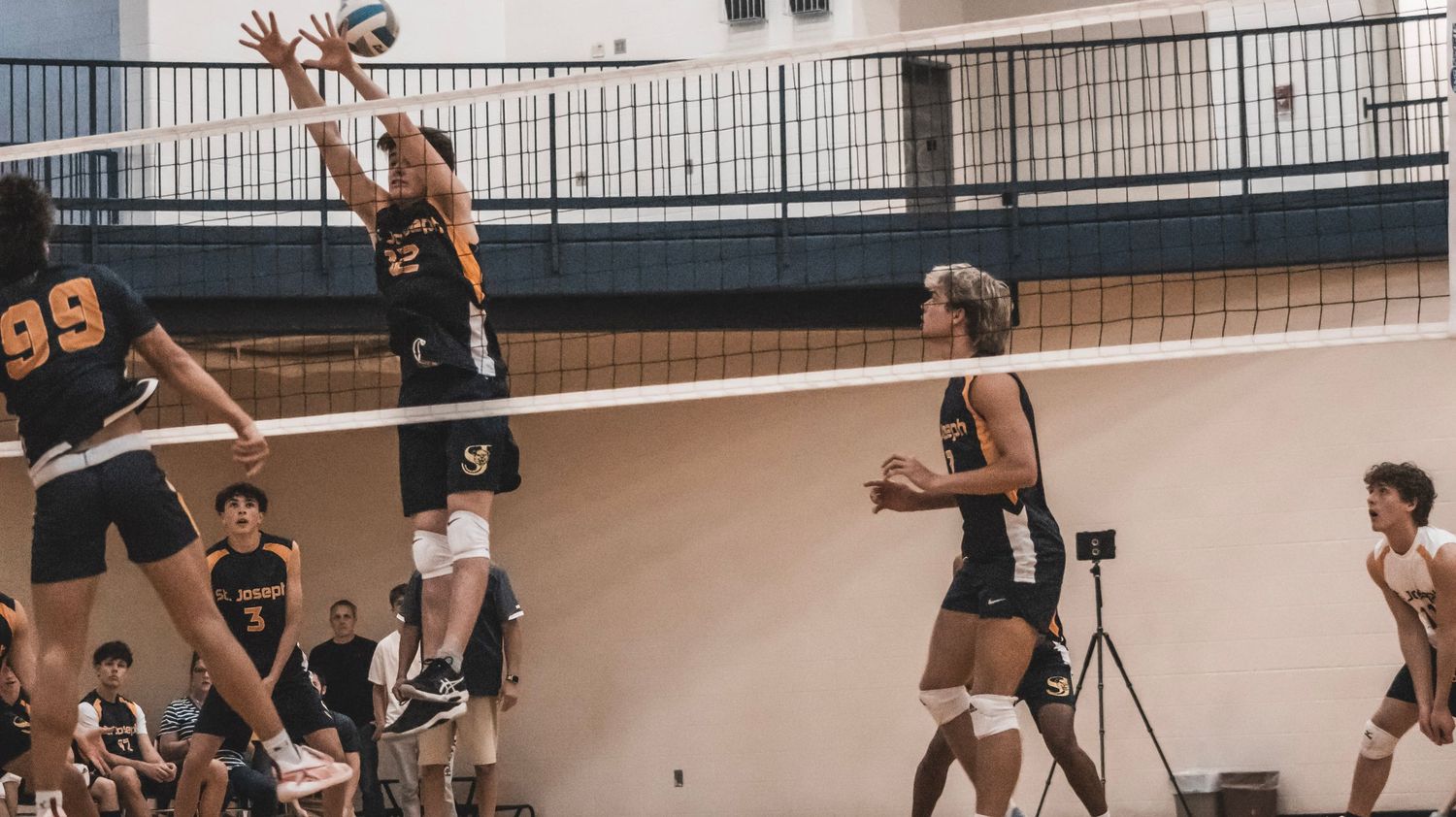 Two players jump to block at the volleyball net in an indoor gym setting.