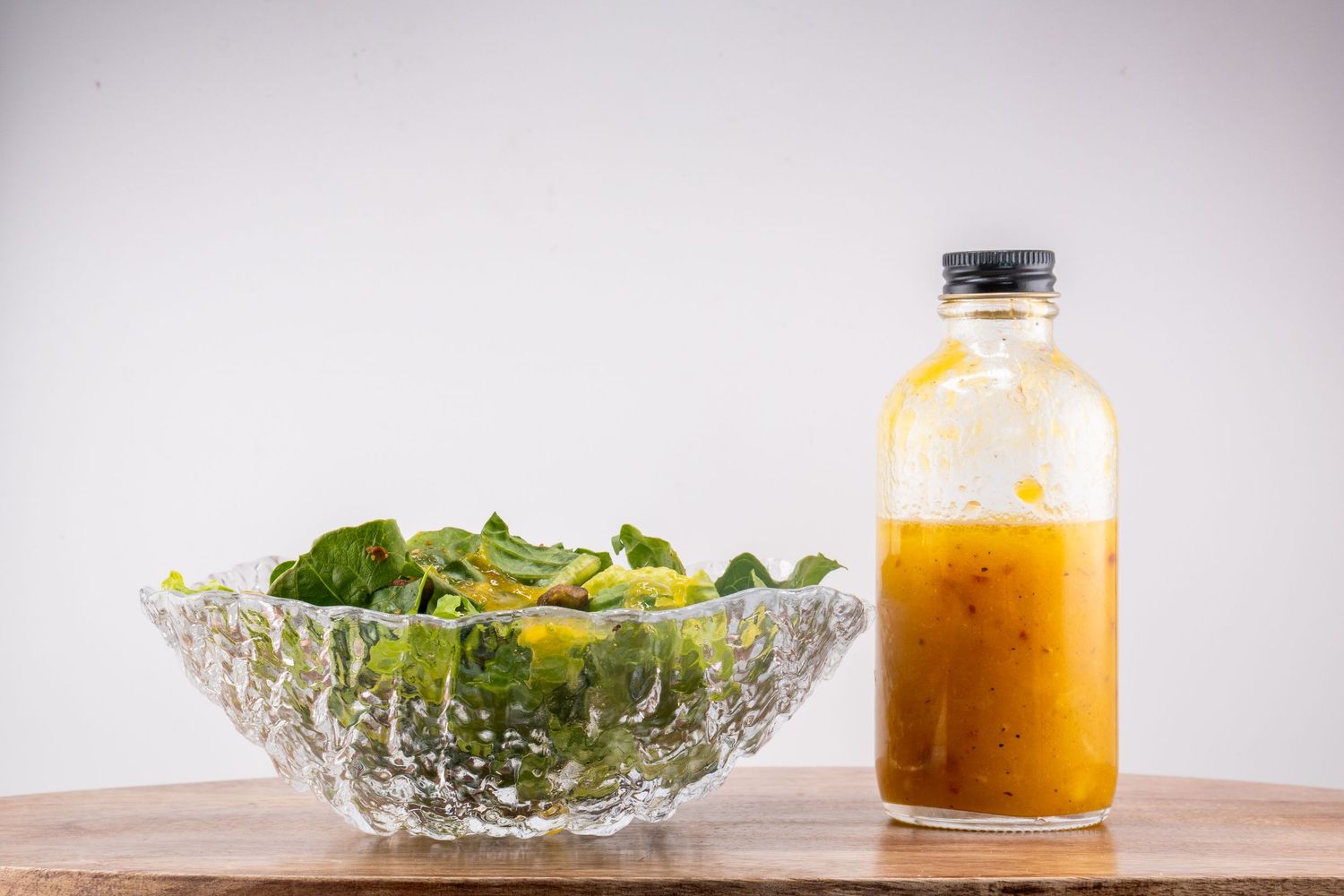 Green salad in crystal bowl with orange dressing bottle on wooden surface.