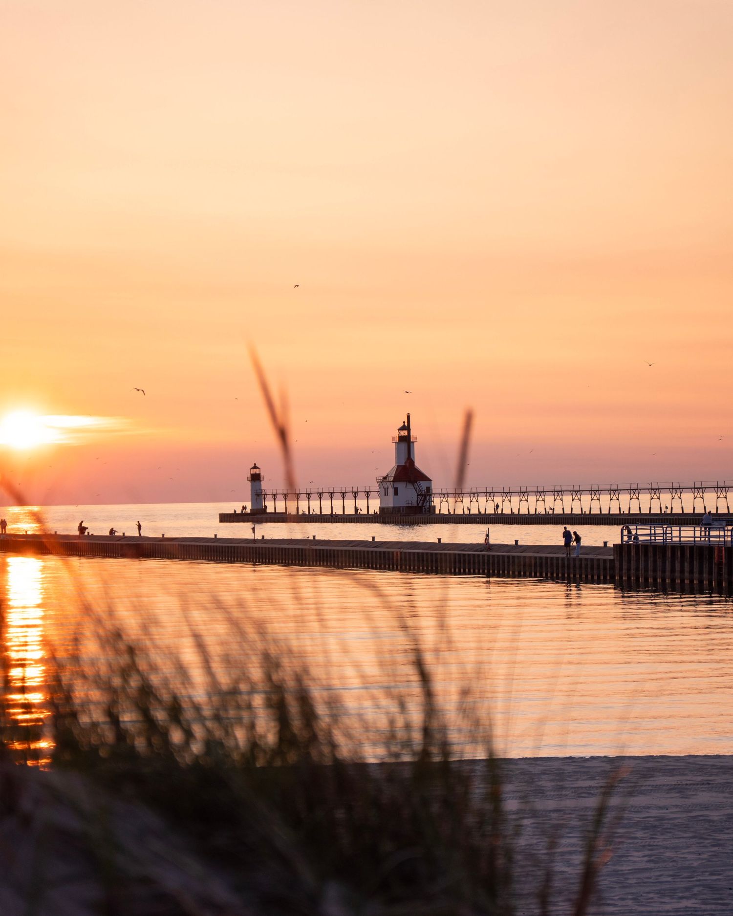 Lighthouse silhouetted against orange sunset sky with reflections on calm harbor water.
