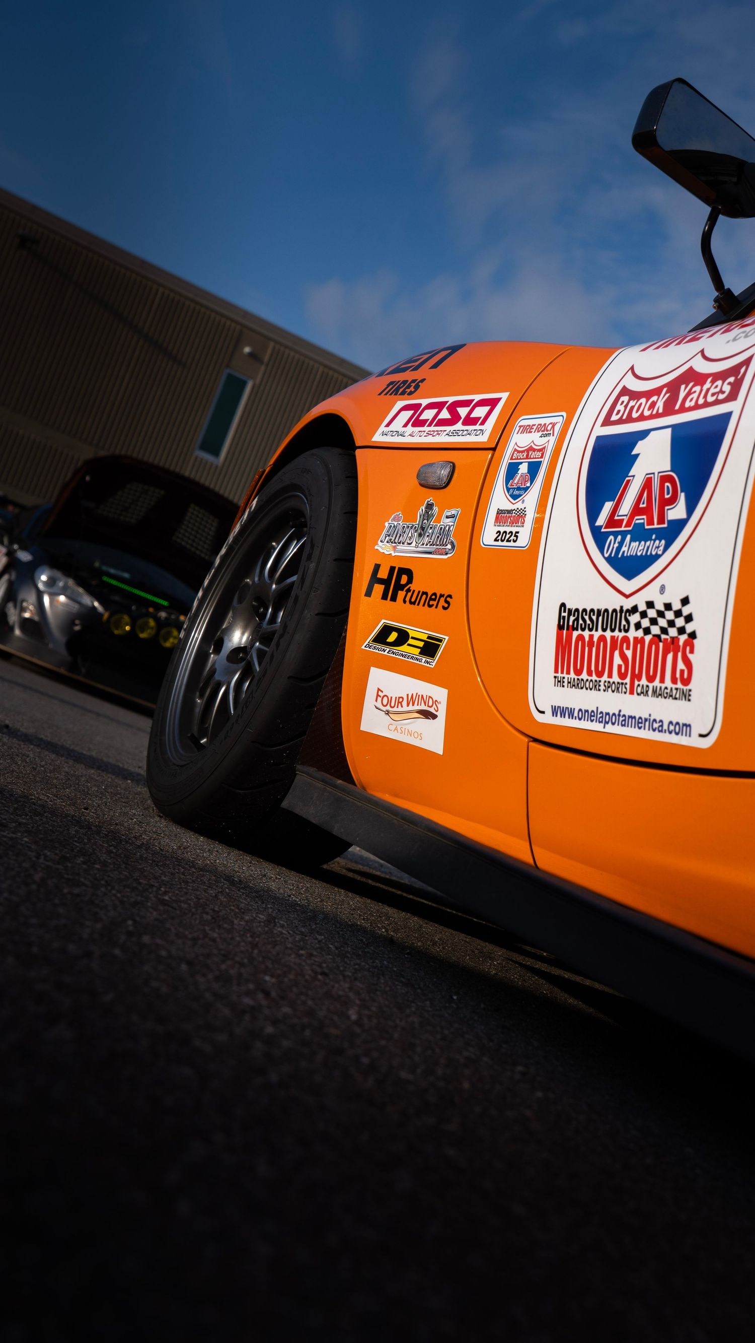 Orange race car with sponsor decals and racing stripes parked in outdoor setting.