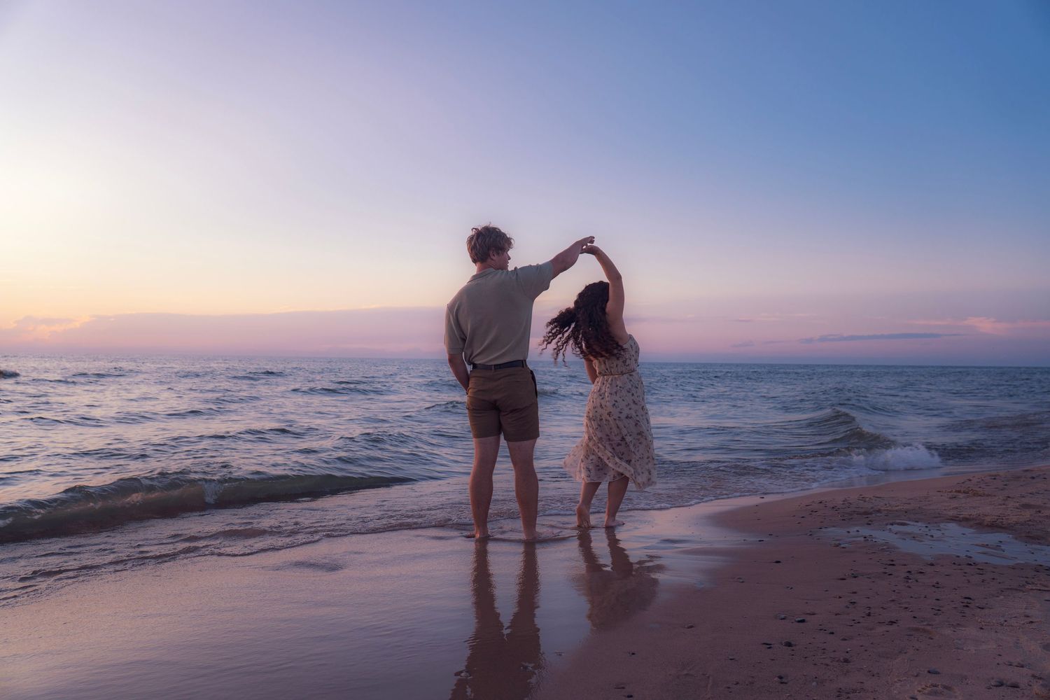 Two people walk along a tranquil beach at sunset, their silhouettes framed against a pastel-colored sky and ocean.