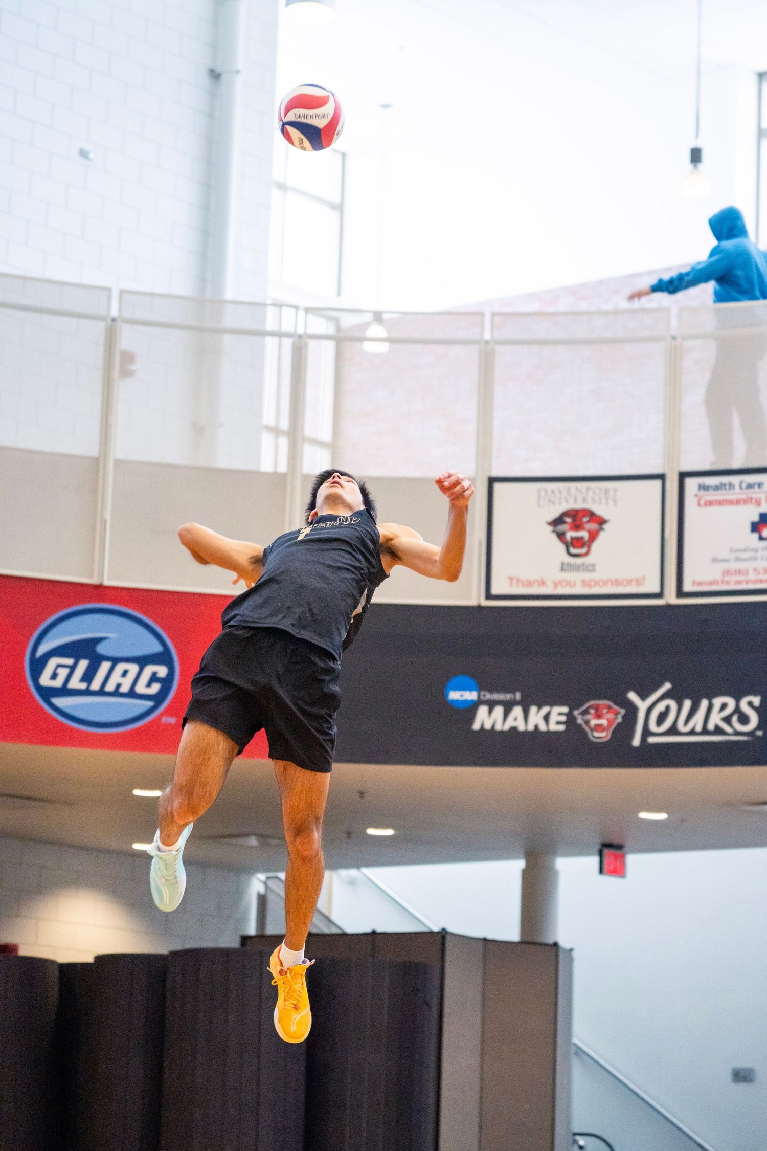 A volleyball player jumps high to spike the ball during an indoor match with GLIAC conference signage visible.