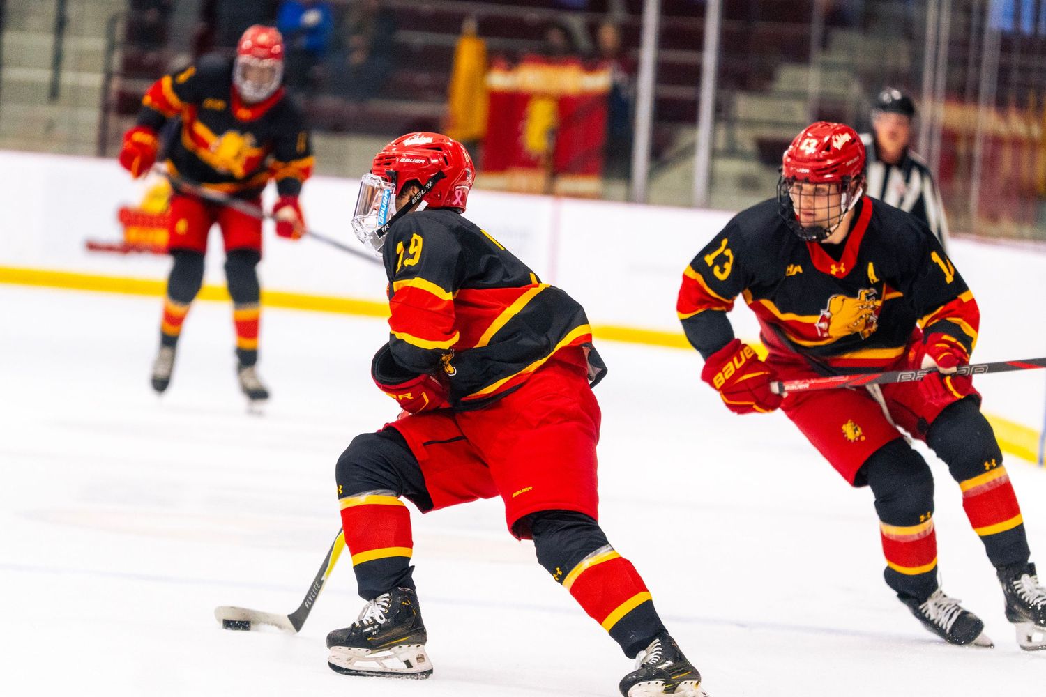 Hockey players in red, black and yellow uniforms skate on the ice during a game, with a referee watching in the background.