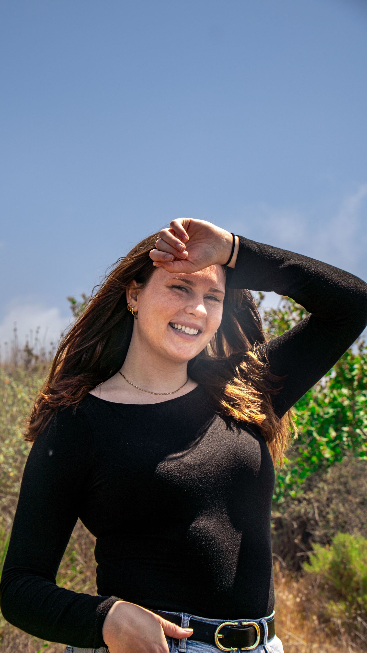 Person in black sweater shielding eyes from sun while posing outdoors on sunny day.