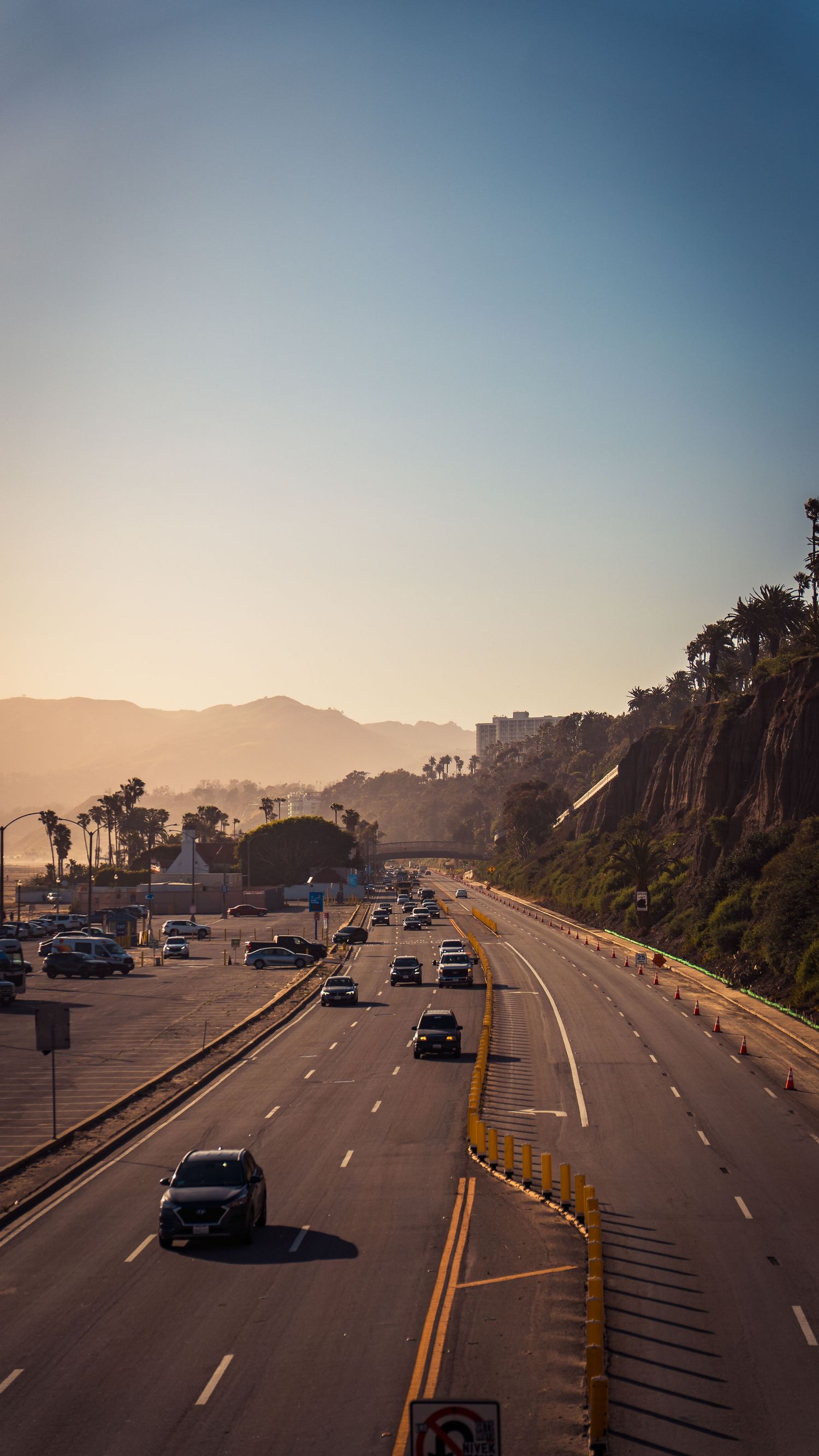 Sun-drenched highway follows coastline with cars traveling between cliffs and ocean.
