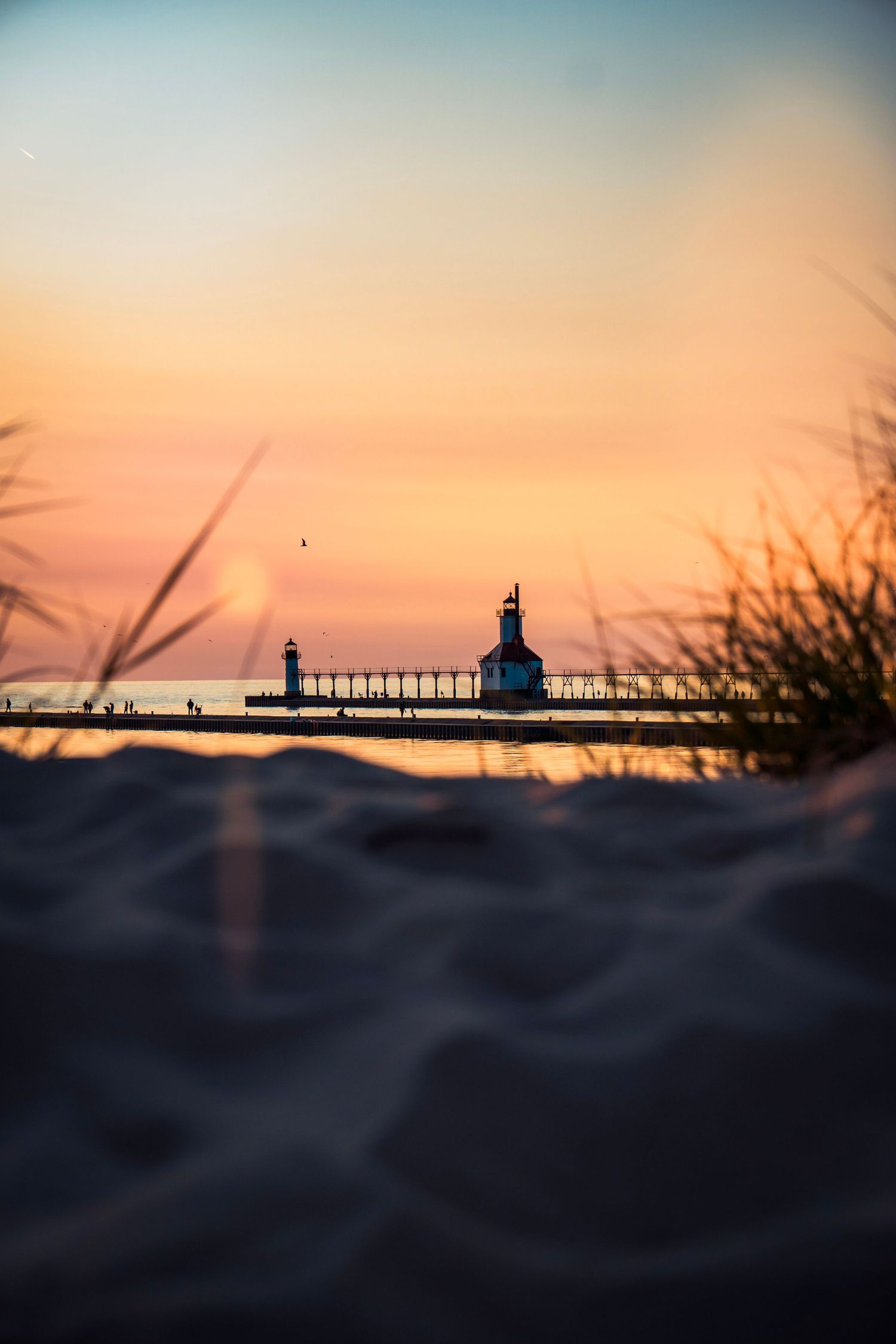 Lighthouse and pier silhouetted against an orange sunset sky, framed by beach grass.