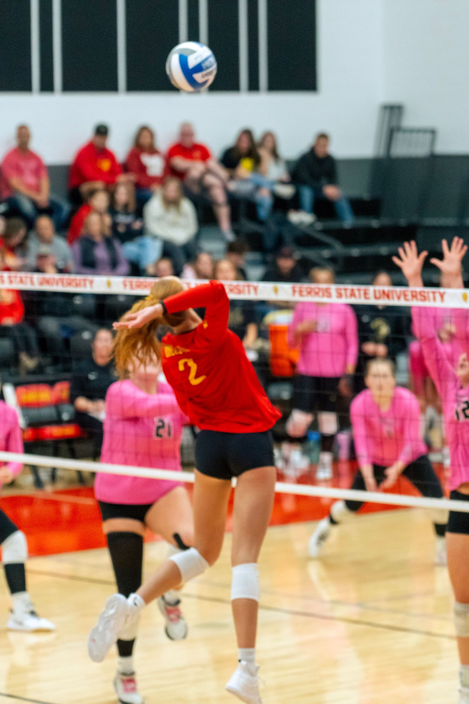Volleyball players in pink and red uniforms dive for a ball during an intense indoor match at a university gymnasium.