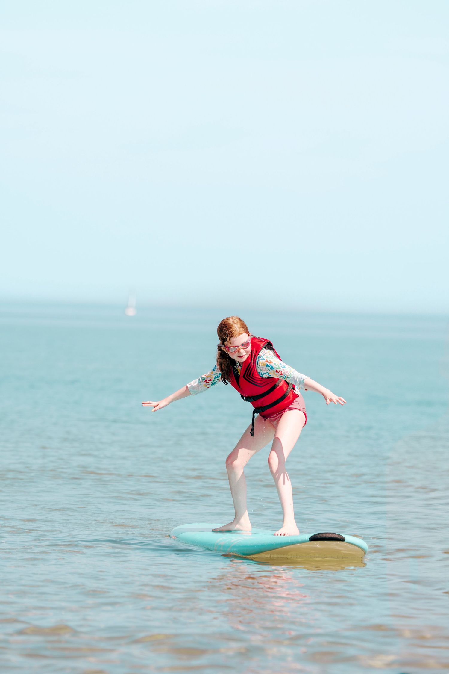 A person in a red shirt practices balancing on a surfboard in shallow turquoise ocean waters.