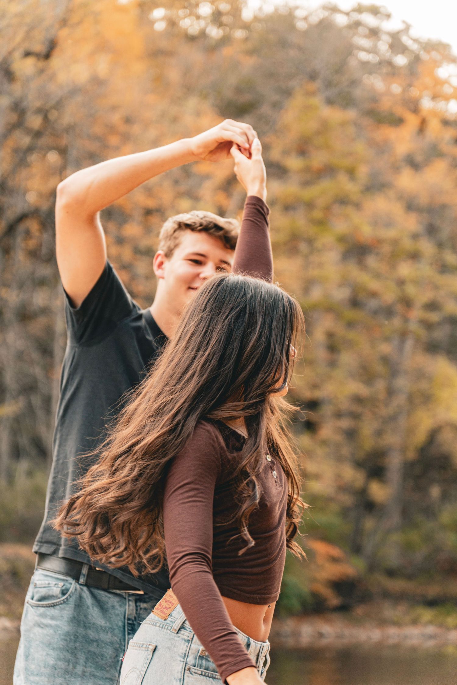 Young couple dances outdoors by a lake as a smiling man twirls a woman with long brown hair beneath vibrant autumn foliage.